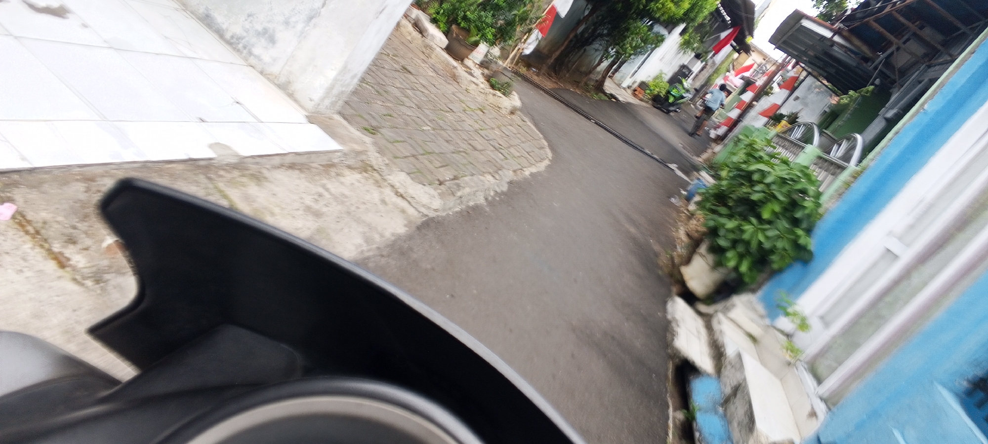 This image, taken from a low perspective, likely from a motorcycle, captures a street scene in Depok, Indonesia. The foreground is dominated by the dark, curved plastic of a motorcycle's front fairing. Beyond this, a paved street curves to the right, bordered by a sidewalk made of interlocking bricks. On the left, a white tiled wall runs parallel to the sidewalk. The right side of the street features a blue building with a white-framed window. Green foliage is visible both in pots and growing along the edge of the street. Several Indonesian flags, red and white, are displayed on poles along the right side of the street, indicating a possible national celebration. In the mid-ground, a person is riding a green motorcycle, and further down the street, another person is riding a bicycle. The scene appears to be an ordinary day in a residential or commercial area.