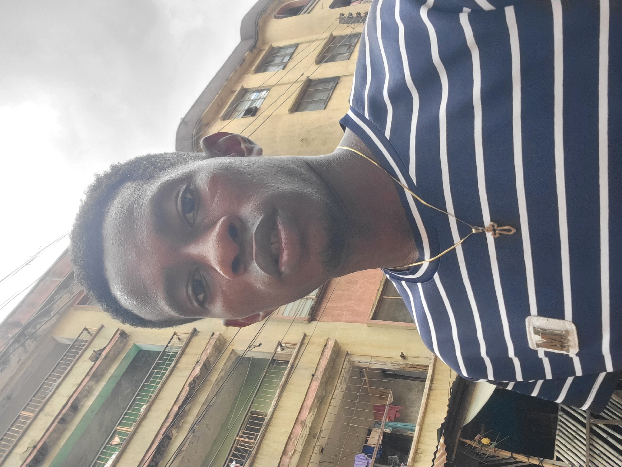 A young man with dark skin and short, dark hair is captured in a selfie. He is wearing a navy blue and white striped t-shirt with a golden cross pendant. The background consists of a weathered, multi-story building with balconies and some laundry hanging outside. The scene appears to be outdoors in a city, with the architecture suggesting a location in Onitsha, Nigeria. The sky is overcast and gray.