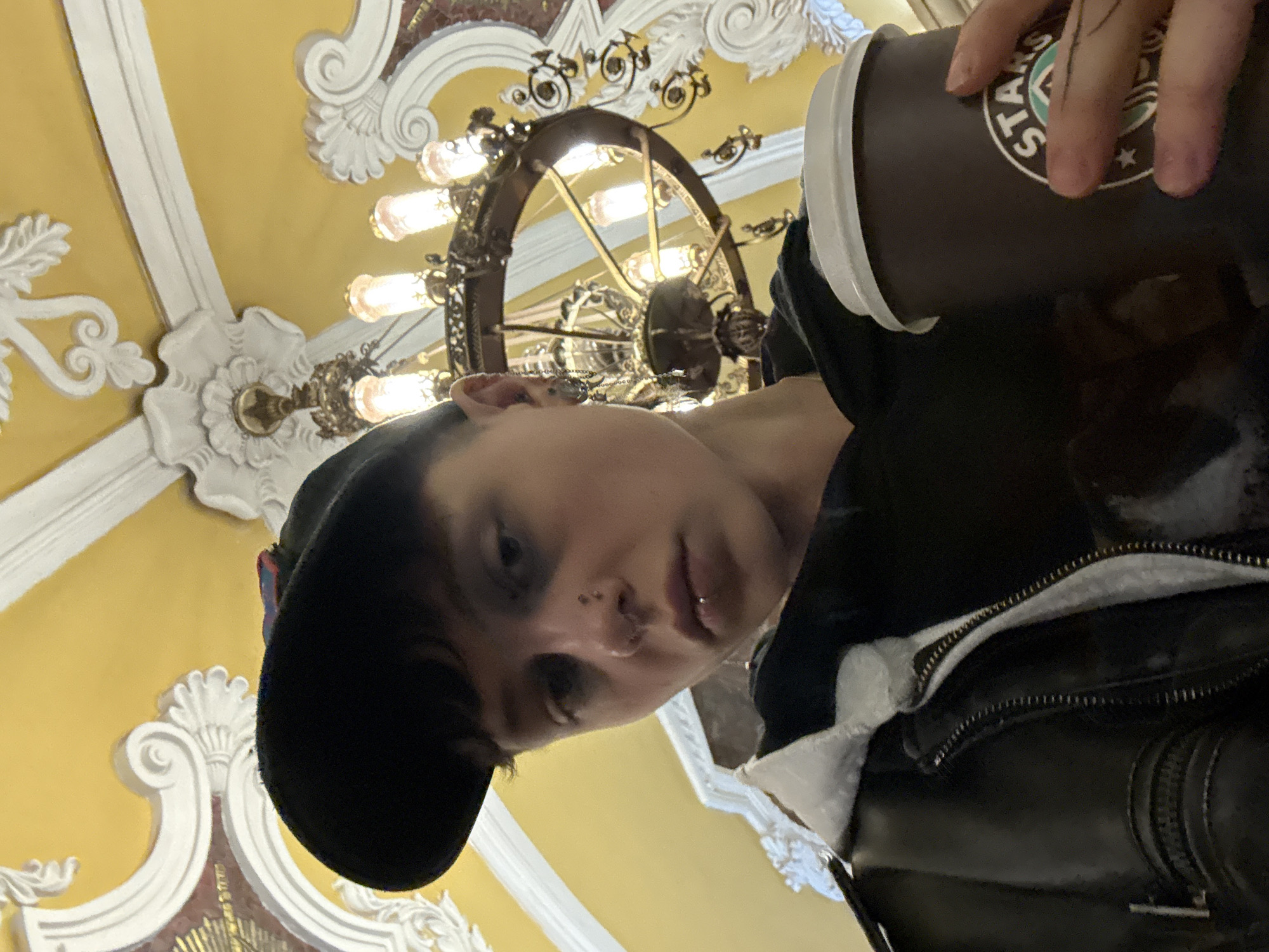 A person is holding a Starbucks cup in what appears to be a subway station in Moscow, Russia. The person is wearing a black cap and a black leather jacket with a zipper. The background features ornate, classical architecture with a large chandelier. The station's ceiling is painted in a yellow hue, adorned with white architectural trim.