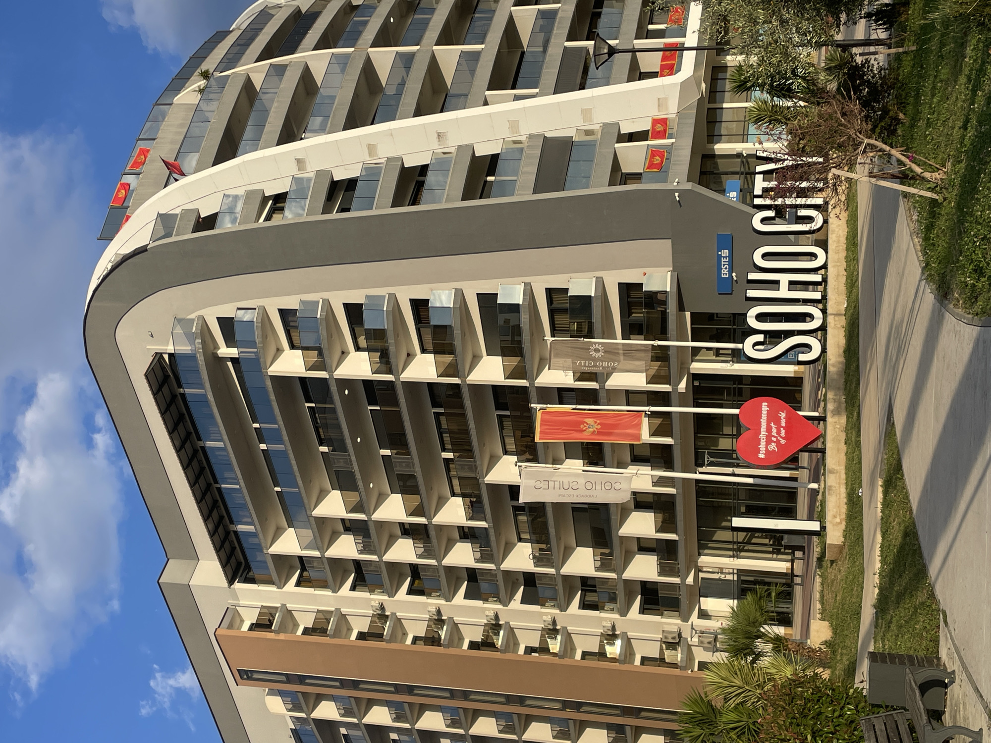 The image depicts the exterior of the Soho City complex in Bar, Montenegro. The building is a tall, modern structure with multiple floors and balconies. It is characterized by its curved facade, a blend of gray, white, and brown colors, and extensive use of glass. The "SOHO CITY" logo is prominently displayed in large, white letters. Several flags of Montenegro are visible along the building's upper floors. In the foreground, there is a large red heart-shaped sign with the hashtag "#sohacitymontenegro" and the phrase "Be a part of our world...". A concrete walkway and some vegetation are also visible.
