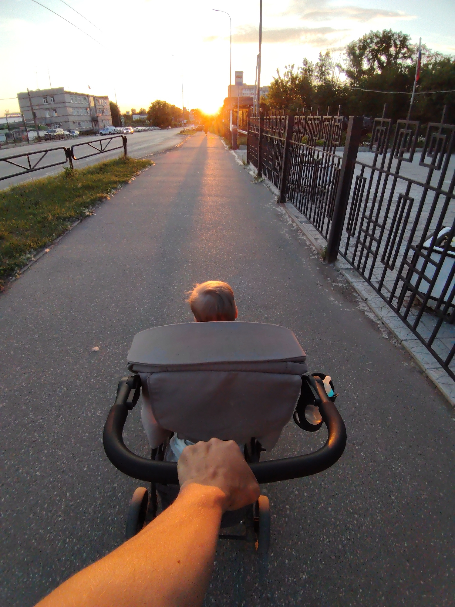 A first-person point-of-view image captures a person pushing a grey stroller along an asphalt sidewalk at either sunrise or sunset. The lower right forearm and hand of the individual pushing the stroller are visible, gripping the black stroller handle. A child with light-colored hair is seated in the stroller, facing away from the camera.
To the left, the asphalt sidewalk is bordered by a narrow grassy strip, beyond which is a simpler black metal fence separating it from a multi-lane asphalt road. Across the road, a grey, multi-story building with multiple windows, characteristic of Soviet-era architecture, is visible in the distance, along with parked and moving vehicles. Overhead wires and streetlights are present on the left.
To the right, the sidewalk is bordered by an elaborate black metal fence featuring distinct geometric, interlocking rectangular patterns. Beyond this fence, dense trees are visible, and a flagpole with a partially visible Russian flag (red, white, and blue horizontal stripes) stands within the treeline.
The sun is low on the horizon, directly ahead, casting a strong, warm orange-yellow glow and long shadows along the entire street, illuminating the pavement and the stroller from the front. The sky is partly cloudy.
This urban scene is located on **Lenin Street (ул. Ленина) in Chaikovsky, Perm Krai, Russia**. The distinctive ornate fence on the right indicates the immediate vicinity of the Chaikovsky City Administration building grounds or the adjacent Park Kultury i Otdykha.