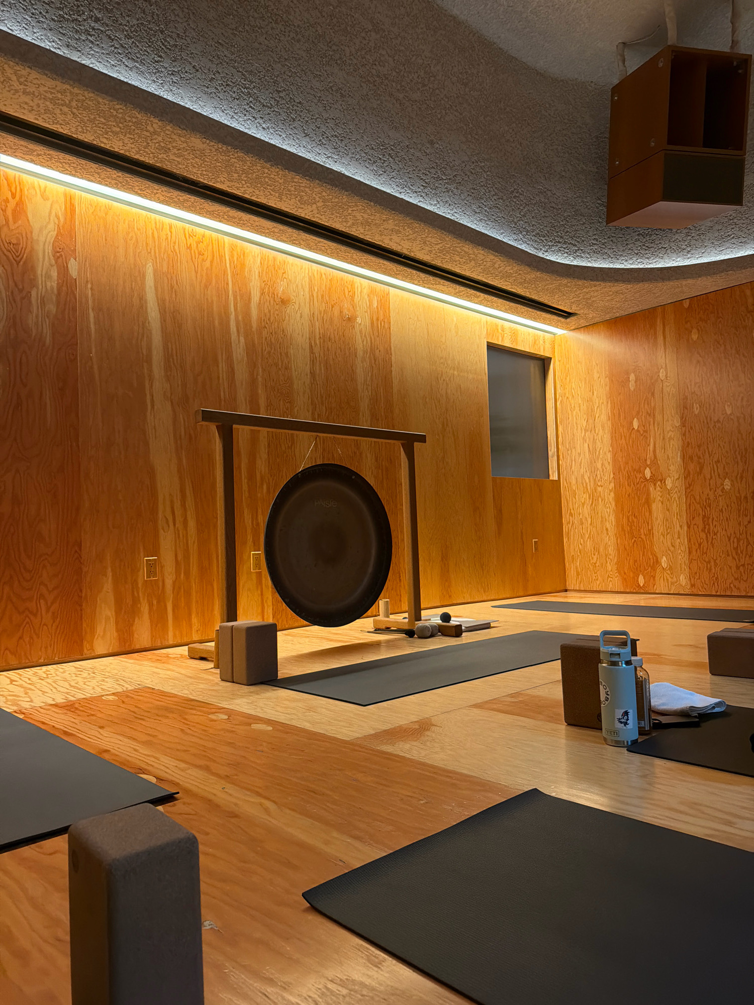 The image depicts the interior of a yoga studio in Los Angeles, California, USA. The room is characterized by its warm, natural wood paneling on the walls and a polished wooden floor. A large gong hangs from a wooden stand in the center of the room, suggesting a focus on sound healing or meditation practices. Several yoga mats are laid out on the floor, indicating the space is prepared for a class. In the foreground, on the right side, a light blue water bottle with a sticker and a small towel are placed near a yoga block. The ceiling features an undulating texture and is illuminated by strip lighting, creating a soft ambiance. A small, dark window is visible on the back wall. The overall impression is one of a serene and minimalist space designed for wellness activities. There are no people present in the image.
