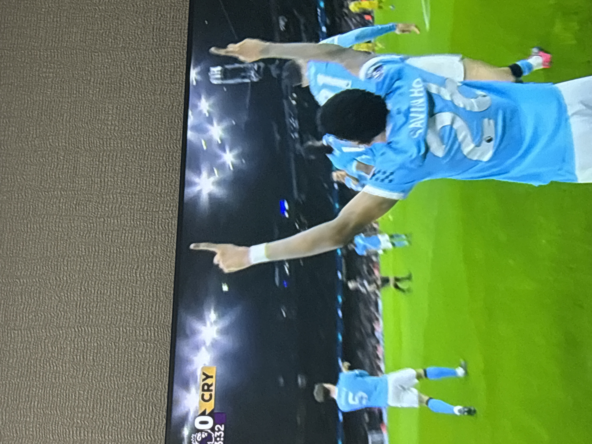 The image shows a soccer game on television, with a close-up of Manchester City players celebrating a goal. The scoreboard indicates "CRY" (Crystal Palace) at 0, with a time of 3:32, likely meaning 3 minutes and 32 seconds into the game. A player wearing number 26, labeled "SAVINHO" is visible. Other players in light blue Manchester City jerseys are nearby, and at least one has his arm raised, pointing upward. The scene is a broadcast of a soccer match, not a physical location in Yerevan, Armenia.