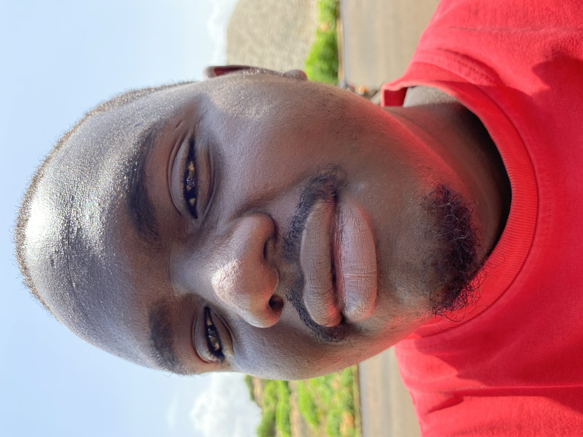 A close-up, head-and-shoulders, outdoor selfie of a Black male taken in Bukuru, Nigeria. The subject is wearing a red t-shirt and has dark skin, a shaved head with a thin hairline, and a small mustache and goatee. The background includes a bright blue sky, some green vegetation on a hillside, and a large pile of gravel. The lighting suggests it is daytime with sunlight casting highlights on the subject's skin.