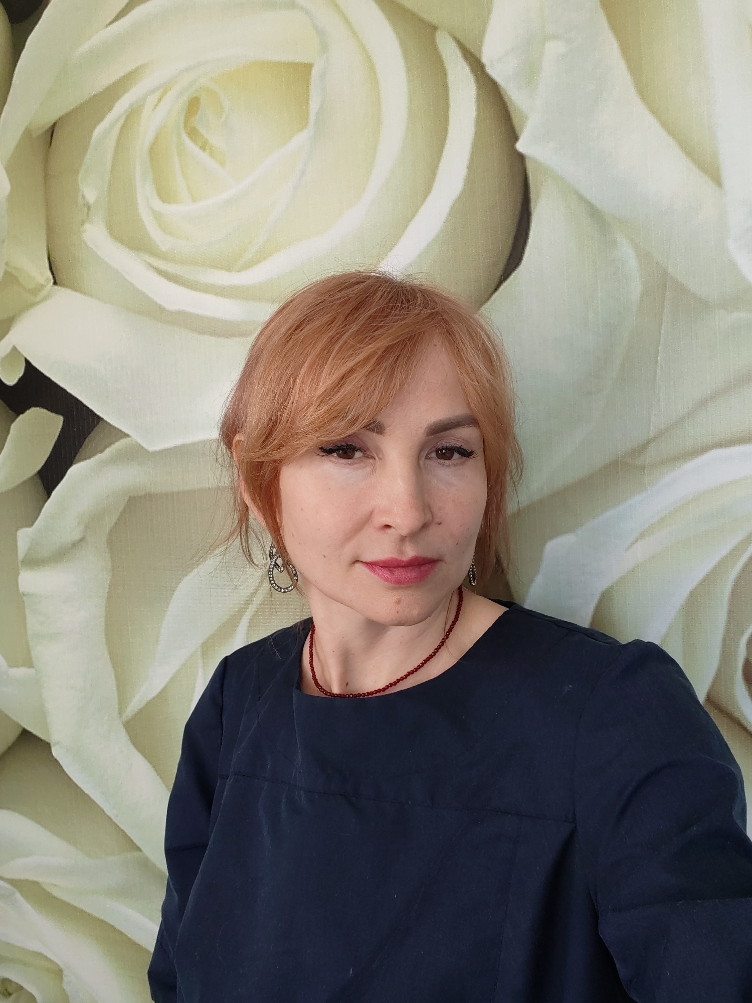 A woman with reddish-orange hair is centered in the frame against a backdrop of large, light beige rose blossoms. She wears a dark blue blouse and a red beaded necklace. She has light skin, reddish lips, and is wearing dangling earrings. The photo appears to be taken indoors, potentially in a decorative or domestic setting. There is no information available to confirm whether the photograph was taken in the city of Chaikovsky, Russia.