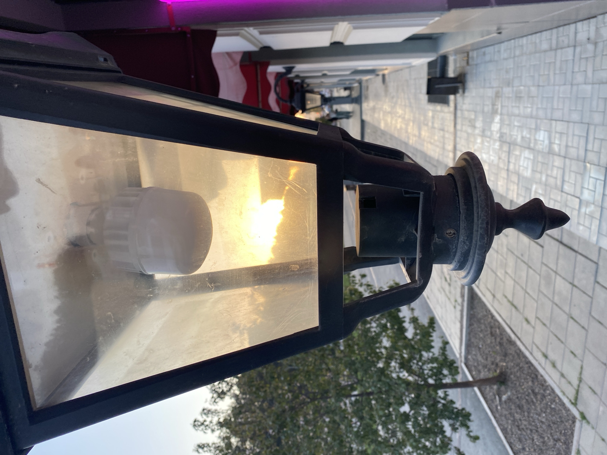 The image is an eye-level, close-up shot of a vintage-style street lamp. The lamp has a black metal frame enclosing a translucent yellowish pane, behind which a bulb is visible. The frame has decorative elements at the top. The background shows a street scene in the Russian city of Yekaterinburg. Paving stones form a pedestrian walkway. A tree with green foliage is to the right. Further down the street, more lamp posts are visible, and a building with awnings is to the left, where a pink light source is noticeable. The street appears to be an outdoor commercial area.