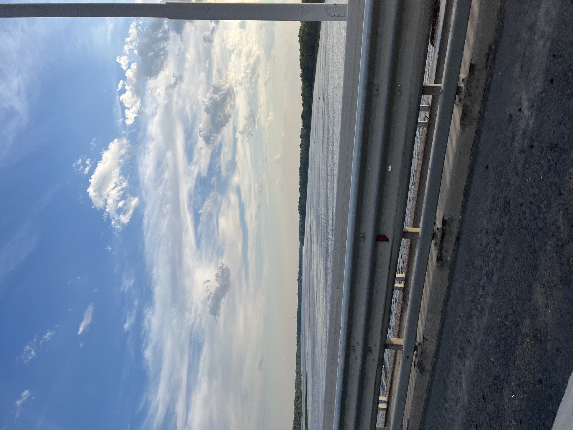 The image depicts a wide, calm river flowing through a landscape bordered by dense green trees. The sky above is a vibrant blue, filled with scattered white and gray clouds. In the foreground, a metal guardrail is visible, suggesting the vantage point is from a bridge or elevated roadway. The road surface in the immediate foreground appears to be asphalt. The scene is likely captured during daylight hours, possibly late afternoon or early evening, given the quality of light. There are no people or specific structures immediately discernible in the midground or background, save for faint outlines of distant structures or possibly boats on the water. The location is identified as Shumeika, Russia.