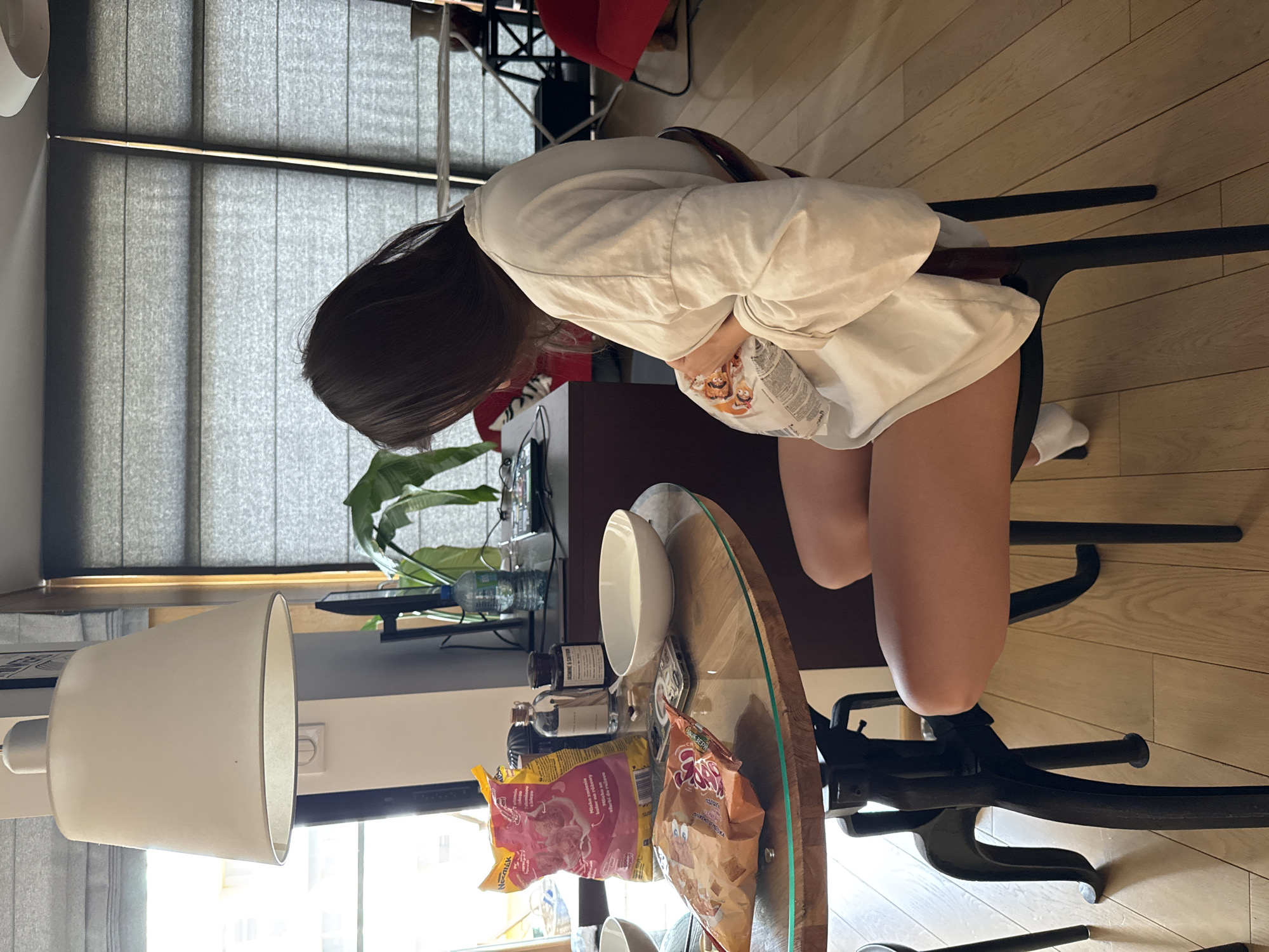 A woman sits at a glass table in what appears to be an apartment in Warsaw, Poland. She is wearing a white sweatshirt, black shorts, and mismatched socks (one white, one black). She is hunched over, holding a bag of chips close to her chest. On the table are a white bowl, various jars and bottles, and several bags of snacks. A lamp is visible in the lower left corner of the image, and a plant is visible on a shelf behind the table. Window blinds are in the background.