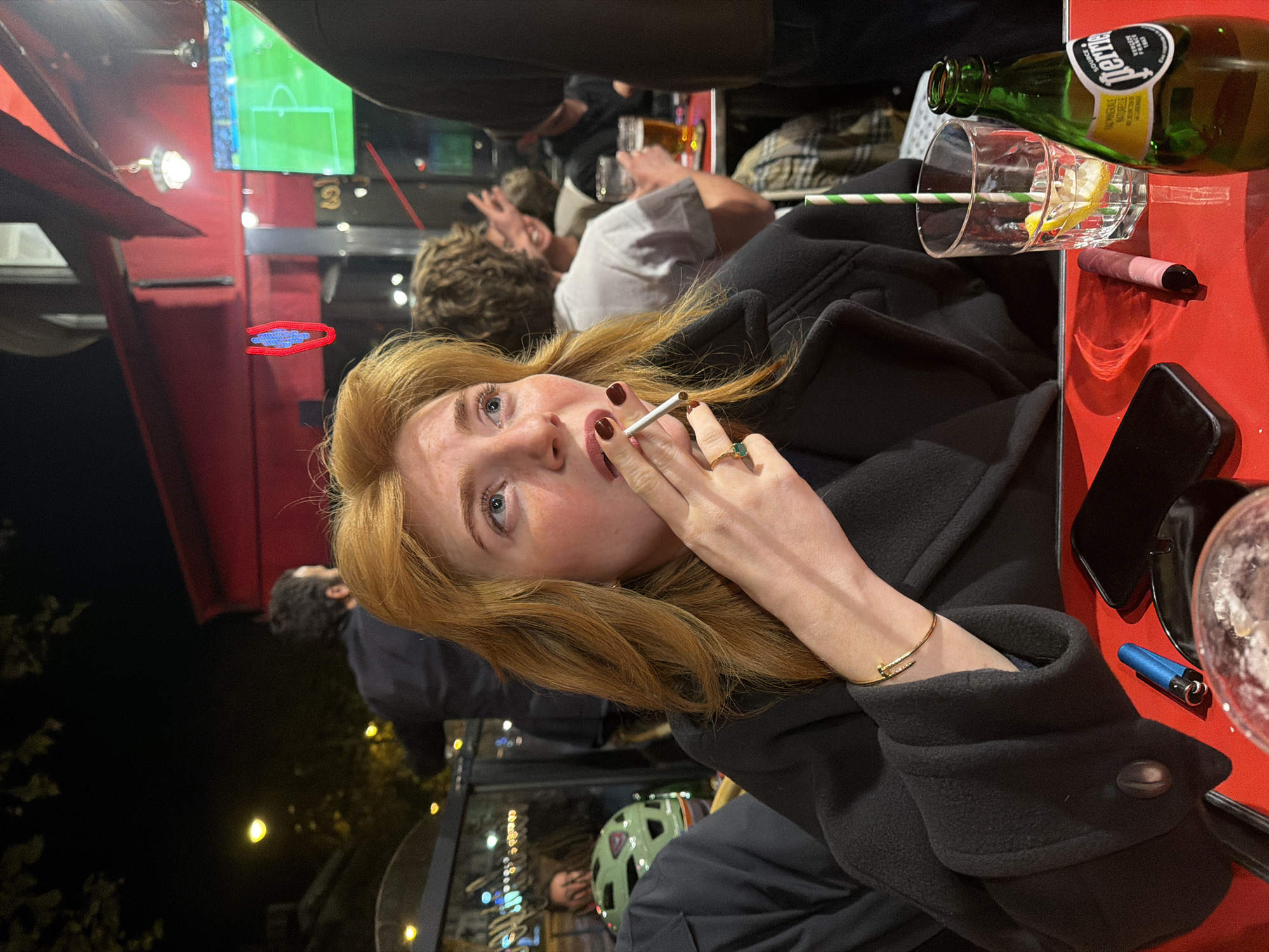 A young woman with red hair is holding a cigarette in her right hand while sitting at a table, likely at an outdoor café in Paris, France. She is wearing a black coat and a gold bracelet. On the table are a glass of water with a straw and lemon, a bottle of Perrier, a pink disposable vape, a black smartphone, and a blue lighter. The table is red. Behind her, a television screen shows a soccer game, and other people are present at the café, including a person in a white shirt making a hand gesture. In the background, lights from buildings and streetlights indicate it is nighttime. A neon sign with a blue edge is visible on the left side. Through a nearby window, there is a bicyclist with a helmet.