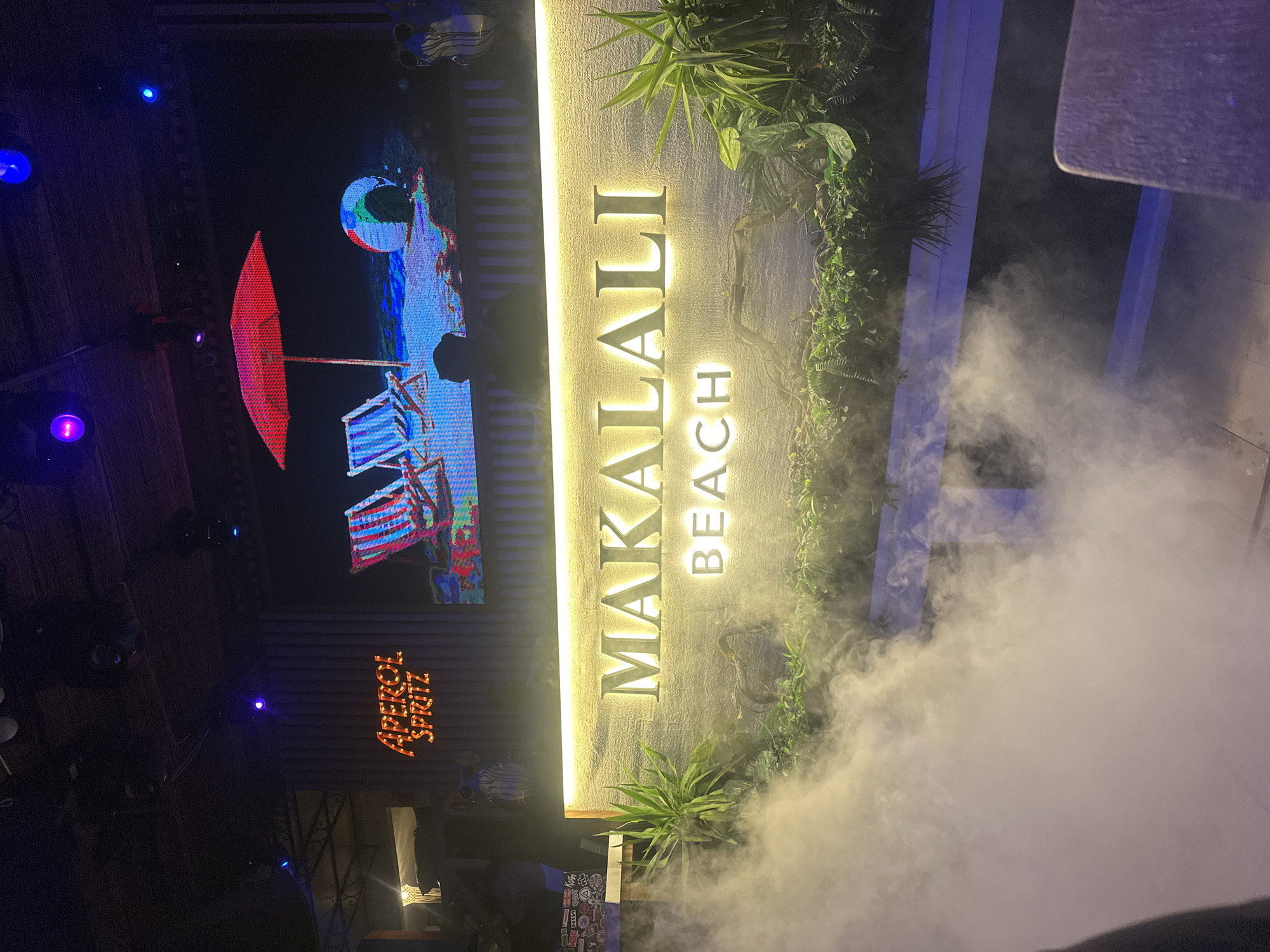 The image depicts an indoor or covered outdoor establishment, likely a bar or restaurant, illuminated primarily by artificial light in a dim environment. Central to the scene is a large, illuminated sign reading "MAKALALI BEACH" in silver-colored, dimensional letters against a textured, light-colored wall, with a warm white strip light above it. Below this sign and extending into the lower foreground, a dense, white artificial fog or smoke billows upwards, partially obscuring the base of the sign and surrounding plants.
Above the "MAKALALI BEACH" sign is a large LED screen displaying a vibrant, stylized image of a beach scene, featuring a red open beach umbrella, two striped beach chairs (one blue, one red), and a colorful beach ball on a blue and white patterned ground. To the left of this screen, a neon-style sign glows orange with the text "APEROL SPRITZ."
Various green plants, some large and leafy, others smaller foliage and ferns, are integrated into the decor along the base of the "MAKALALI BEACH" sign, partially enveloped by the fog. Dark, thin branches are visible within this greenery. The ceiling above is dark, possibly thatched or planked, with multiple spotlights casting blue and purple light. Structural elements, including metalwork, are visible in the upper left. Decorative, stylized animal figures (one dark, one blue) are mounted on the wall to the left and right of the LED screen. In the lower right corner, the edge of a light-colored table is visible. No discernible people are present in the image. The location is Unknown, Unknown.