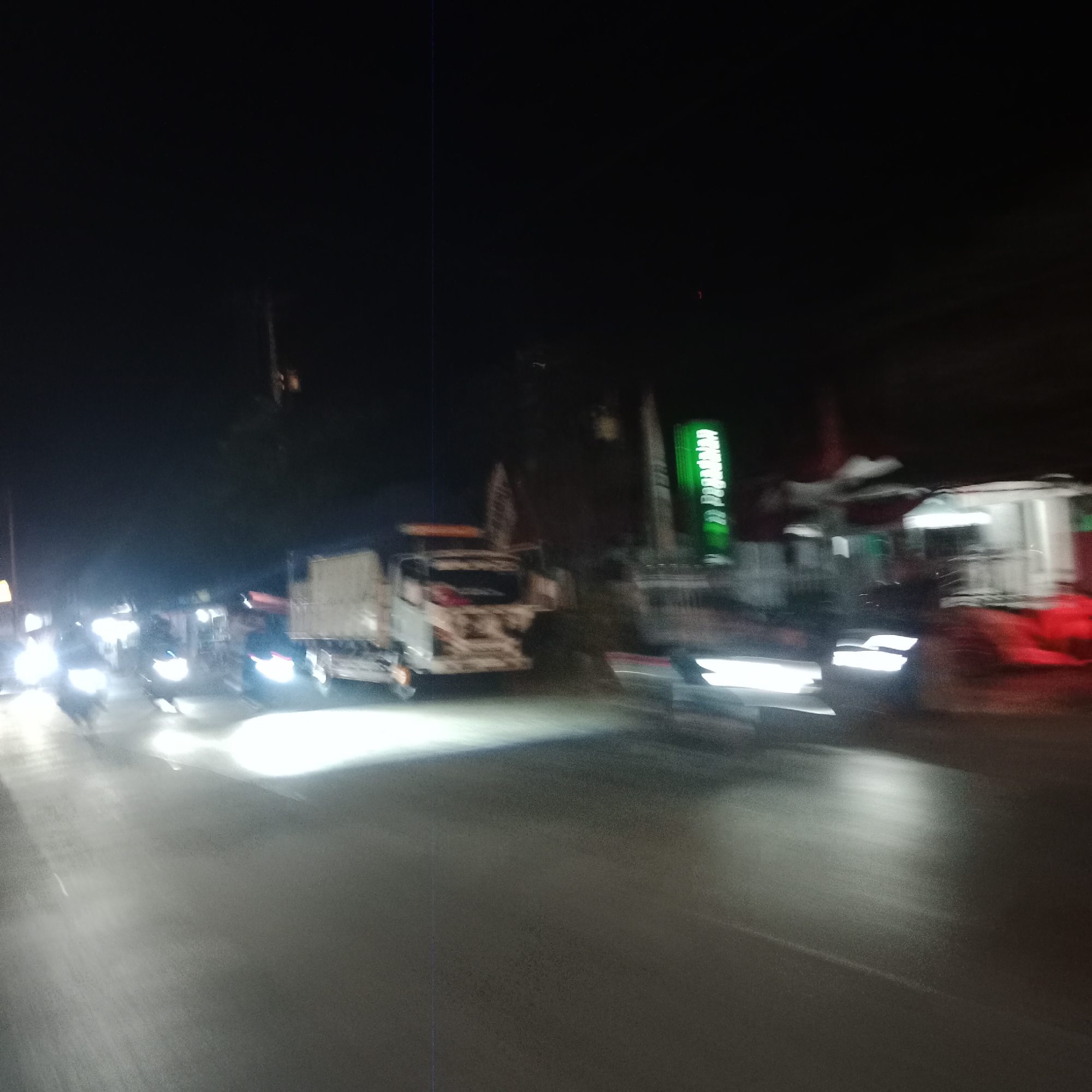 The image depicts a street scene at night in Pilang Kidul, Indonesia. A white truck is visible in the center-left of the frame, appearing to be in motion. In the foreground, several motorcycles are also moving, indicated by their bright headlights. To the right of the truck, a building with a vertical green sign is present, and further to the right, another building with white siding and red accents is visible. The road surface is dark and wet, reflecting the lights from the vehicles and buildings. The overall impression is of a busy, illuminated street at night.