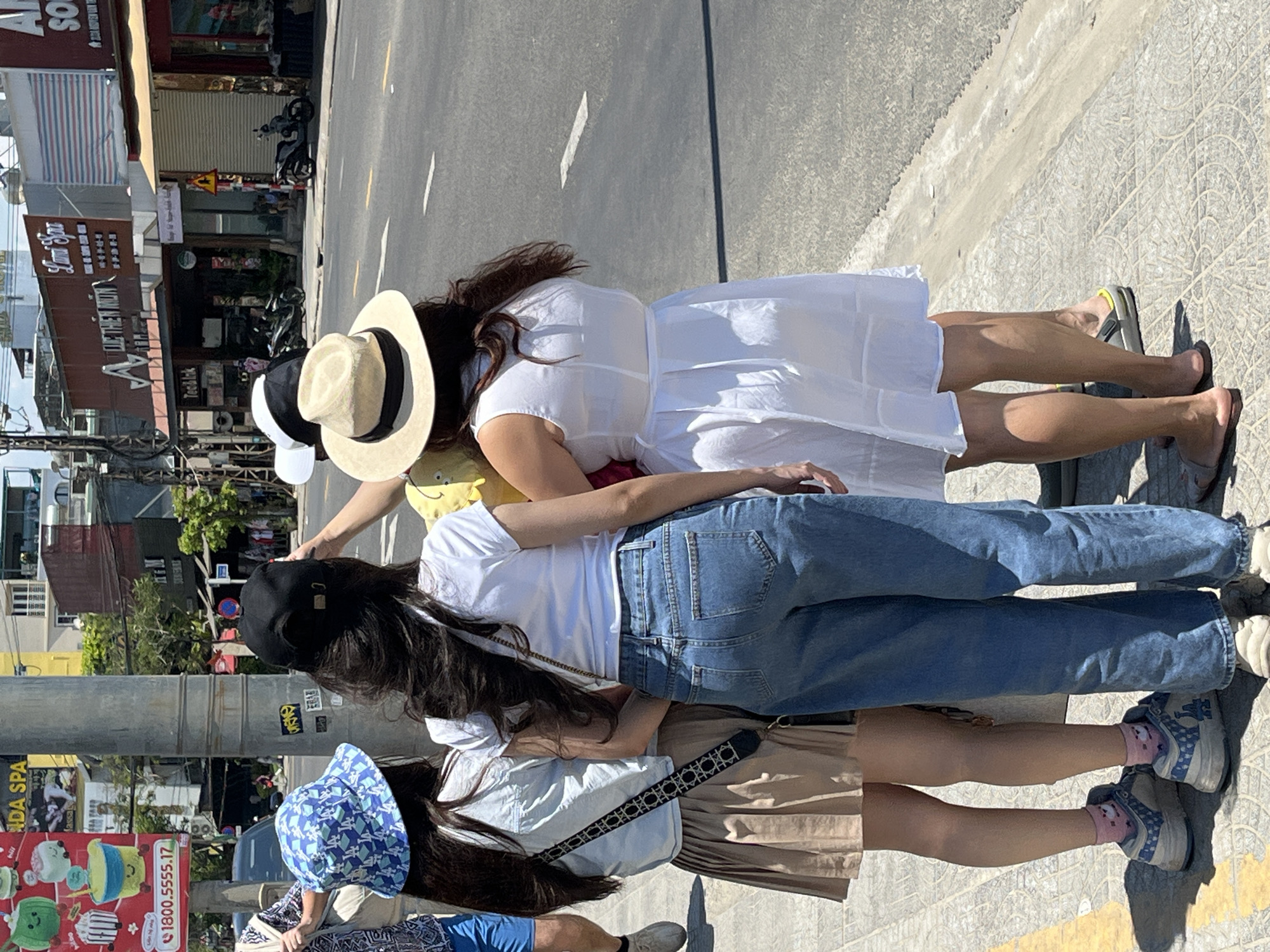 Here's a description of the image:
The image shows a group of four women standing on a sidewalk in Đà Nẵng, Vietnam. From left to right: A woman wearing a blue patterned sun hat, a light blue shirt, and a blue patterned short sleeve shirt. The next woman is wearing a black baseball cap, a white t-shirt, blue jeans, and sneakers. The third woman wears a yellow sundress, a sun hat, and a white dress. The fourth woman is wearing sandals. A road and buildings are visible in the background. A sign with "1800.5555.17" and "ADA SPA" is visible. "Tea & Ink" and "LUC THIÊN MON" are also visible.