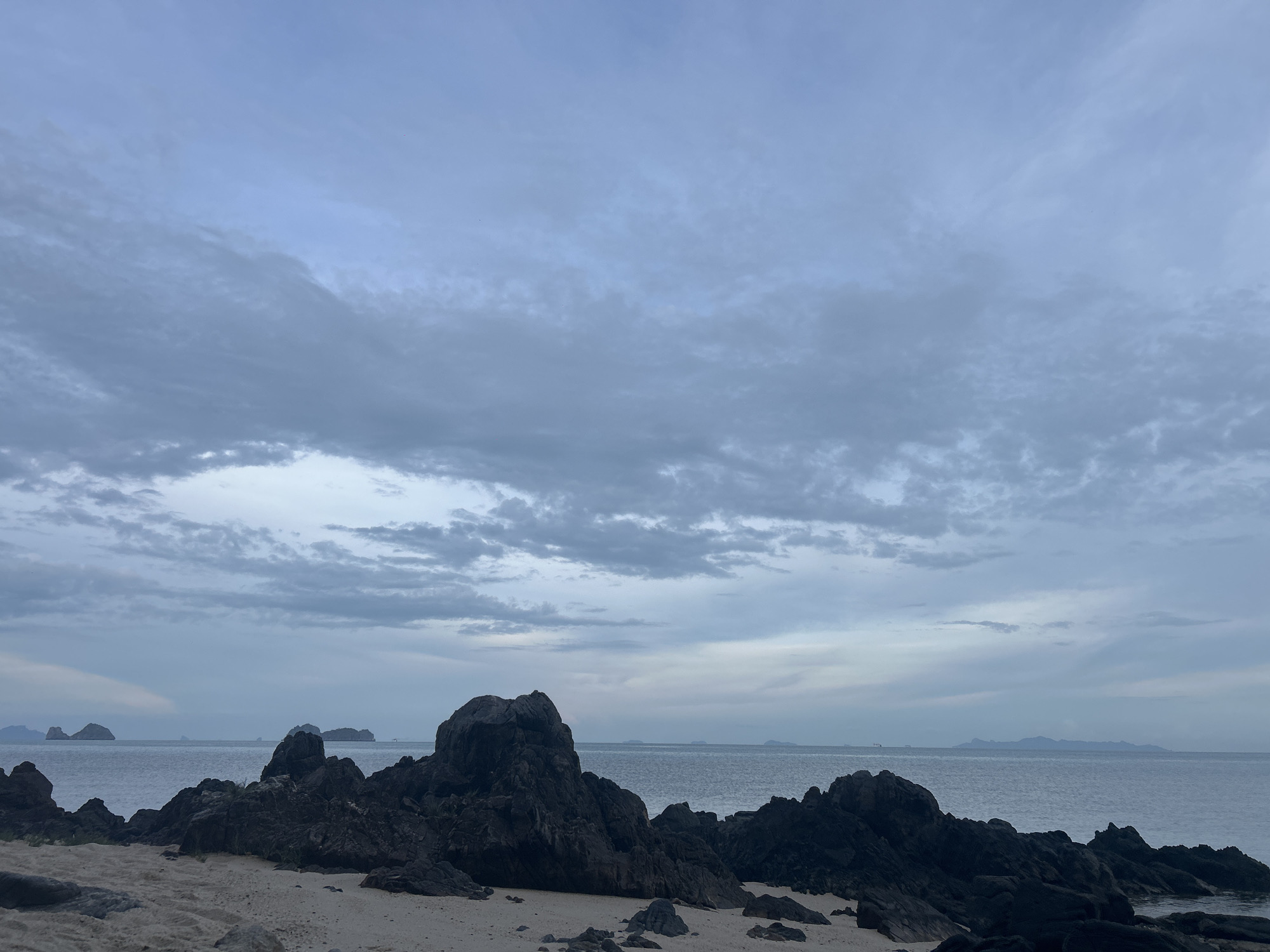 The image depicts a coastal scene, likely near Baan Taling Ngam, Thailand, characterized by a rocky shoreline with dark, irregular rock formations scattered along a sandy beach. The foreground consists of light-colored sand mixed with smaller rocks. In the midground, the ocean extends to the horizon, with several small islands or landmasses visible in the distance. The sky is overcast with a dense layer of grey clouds, creating a muted, somber atmosphere. There are no people visible in the image. The scene overall is natural and unpopulated.