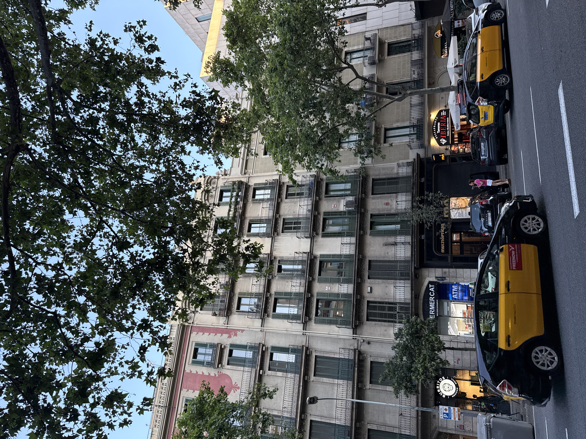 The image depicts a street scene in Barcelona, Spain, characterized by traditional European architecture and bustling city life. In the foreground, a black and yellow taxi, identified by the "FREENOW" logo, drives on the right side of the road. Behind the taxi, several other vehicles are parked or driving, including a black car with a Spanish license plate. The buildings lining the street are multi-story structures with balconies, shuttered windows, and various shopfronts on the ground floor. One shop prominently displays "BURRITOS MEXICAN GRILL" and "SUPERMERCAT," while another advertises "RESTAURANTE TABERNA IBERIA." A few people are visible on the sidewalk, including a woman in a pink top and white shorts standing near a parked car and another individual near the entrance of a shop. Trees with lush green foliage are interspersed along the street, providing shade and adding to the urban landscape. The sky above is a clear blue, suggesting fair weather.