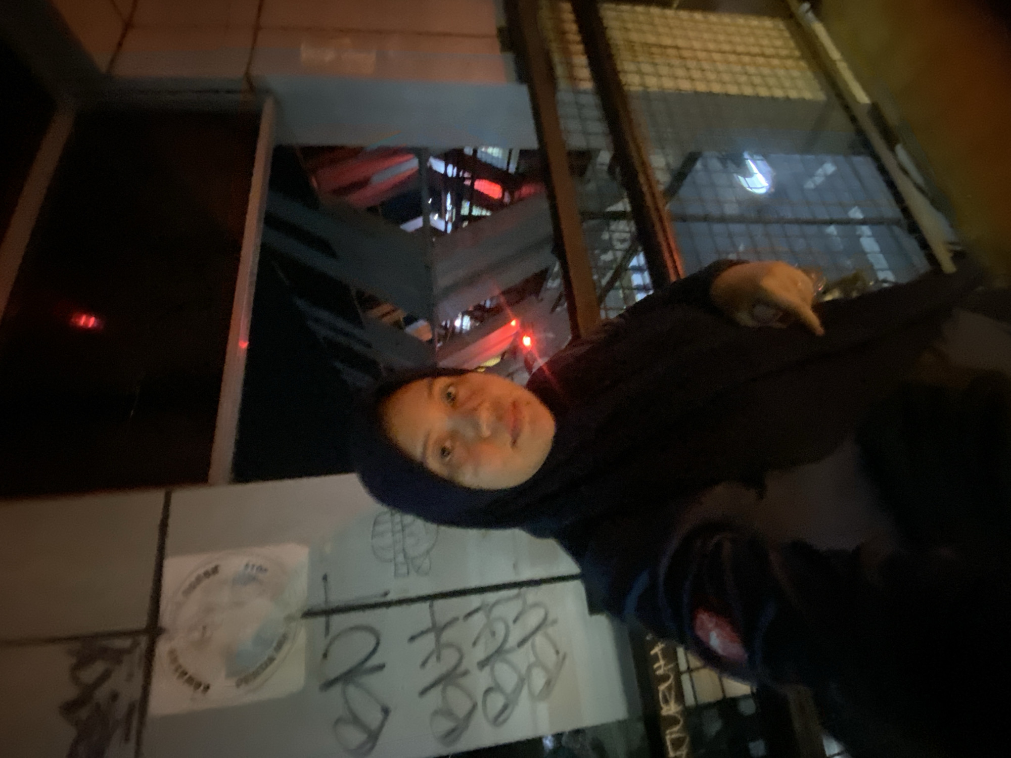 A woman wearing a black hijab and jacket is taking a selfie at night. She is standing on an elevated walkway with a metal railing and a wire mesh fence. Behind her, there is a gray wall with graffiti on it, including some circular text. Further in the background, there is structural metalwork, some of which is illuminated by red lights. The woman is looking at the camera and her right hand is visible, with her index finger extended as if pointing. The overall environment suggests an urban setting, possibly a pedestrian bridge or overpass. The provided information indicates the location is the city of Banjarbaru, Indonesia, though this specific context isn't visually evident in the image itself.