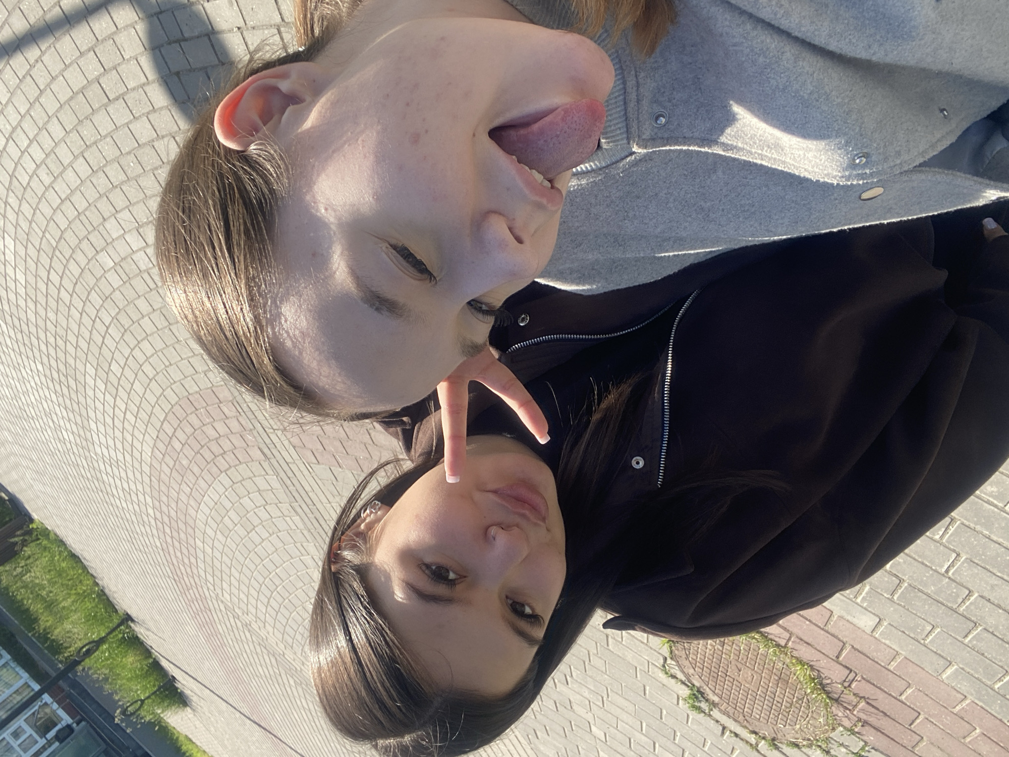 Two young women pose outdoors in Chaikovsky, Russia. One woman with light skin and brown hair makes a face, sticking out her tongue. She wears a light gray jacket. The other woman, with dark hair and a brown jacket, holds up two fingers in a peace sign near her face. They are standing on a paved area with small, square blocks. A patch of green grass and a building are visible in the background. A manhole cover is partially visible on the ground.