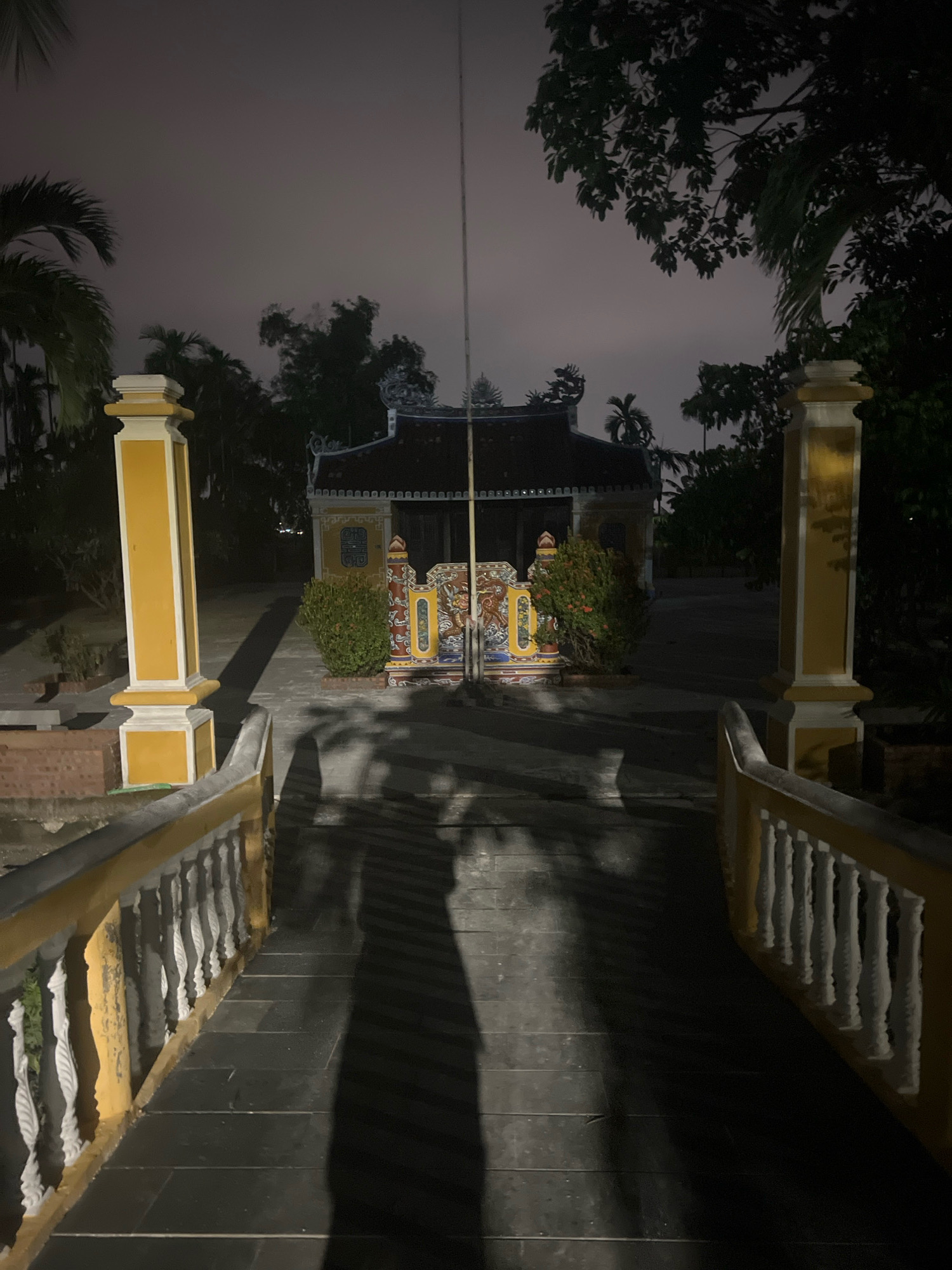 The image shows a nighttime view of a courtyard leading to a traditional building, likely a temple or ancestral house, in Hoi An, Vietnam. The scene is dominated by the silhouette of the building against a dimly lit sky. In the foreground, a stone pathway is lined with ornate yellow and white balustrades, casting long shadows from unseen light sources. Two prominent yellow pillars, each topped with a white cap, stand on either side of the pathway, flanking the entrance to the courtyard. The building itself features a dark, tiled roof with decorative eaves. In front of the building, there is a vibrant, multi-colored ornamental screen or gate with intricate carvings and a small, red-flowering bush to its right. Trees and foliage are visible in the background, adding to the tropical ambiance. The overall impression is one of serene, historical architecture under the cover of night.