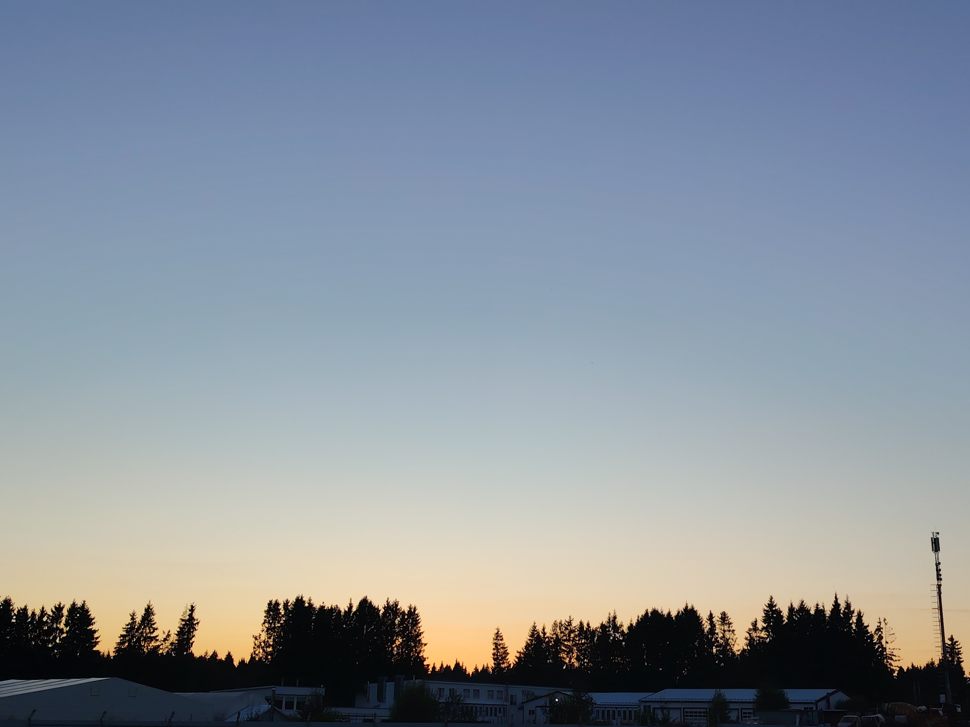 The image captures a silhouetted landscape of trees and buildings against a gradient sky, transitioning from a pale blue at the top to a warm yellow-orange near the horizon. The scene appears to be set in the city of Khorugvino, Russia, likely during dusk or dawn. In the foreground, the tops of several low-rise buildings are visible, some with white roofs. A dense line of dark evergreen trees forms a silhouette across the middle ground. To the right of the frame, a tall telecommunications tower with visible antennas stands prominently against the sky. The sky itself is clear, with only faint wisps of clouds and a few small, distant birds. The overall impression is one of a quiet, rural or semi-rural environment at the edge of a populated area.