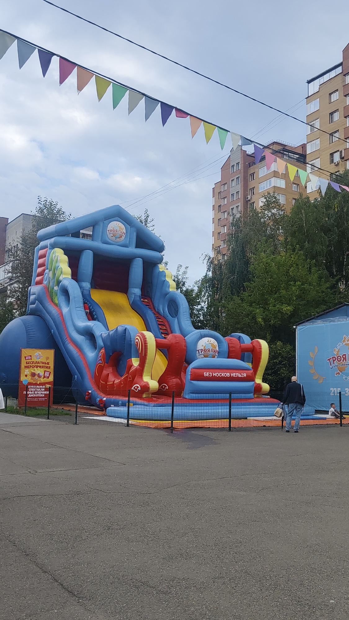 The image depicts a large, inflatable children's playground, predominantly blue, yellow, and red, with a central slide. The playground is set up outdoors, with a paved ground in the foreground and apartment buildings visible in the background. Colorful triangular flags are strung across wires above the playground. A sign with Cyrillic text, which translates to "Free Events" and "TROY PARK," is posted to the left of the inflatable structure. Another sign on the inflatable itself reads "NO SOCKS ALLOWED." A few people are visible in the distance, including a man walking away from the camera and a child sitting on a bench. The overall scene suggests a public recreational area in an urban environment. The text on the signs indicates that this location is in Krasnoyarsk, Russia, specifically at "TROY PARK" entertainment center.