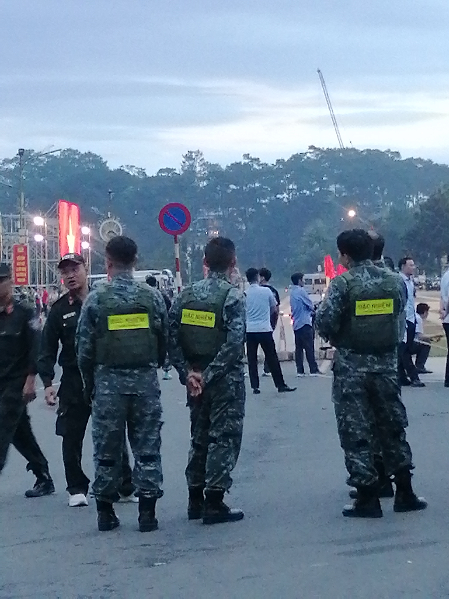 This media file shows a group of people, seemingly law enforcement or security personnel, in what appears to be an outdoor public space in Da Lat, Vietnam. The subjects are primarily men, many wearing camouflage uniforms and tactical vests. Some of the vests have bright yellow rectangular patches with text on them, which appears to be Vietnamese. Specifically, "BẢO NIỆM" is visible on multiple vests.
The scene is set during twilight, with a soft, overcast sky. In the background, trees line a hill, and a construction crane is visible against the sky. There are also lights and structures that suggest an event or gathering is taking place, possibly a festival or a public assembly. The ground appears to be asphalt.
The individuals are standing, some facing away from the camera, while others are engaged in conversation or observing their surroundings. The overall mood seems to be one of vigilance and readiness, consistent with a security detail. There are no strong emotional cues discernible from the individuals' postures or expressions, but their attire and formation suggest a functional purpose. A traffic sign with a red circle and a blue center is also visible on a pole, indicating a restricted area or rule.