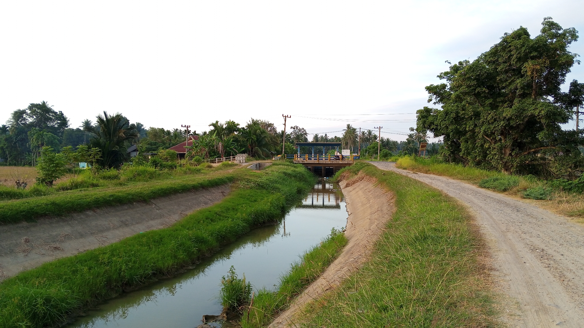 The image depicts a rural scene in Uteunreutoh, Indonesia, featuring a concrete canal with grassy banks running through the center. A dirt road with sparse vegetation parallels the canal on the right. In the distance, a small structure with a blue roof spans the canal, possibly a bridge or water gate. A person on a motorcycle is visible on this structure. The background is filled with lush green trees, some palm trees, and a few houses with red roofs. Overhead power lines are also present. The sky is mostly clear with some overcast clouds.