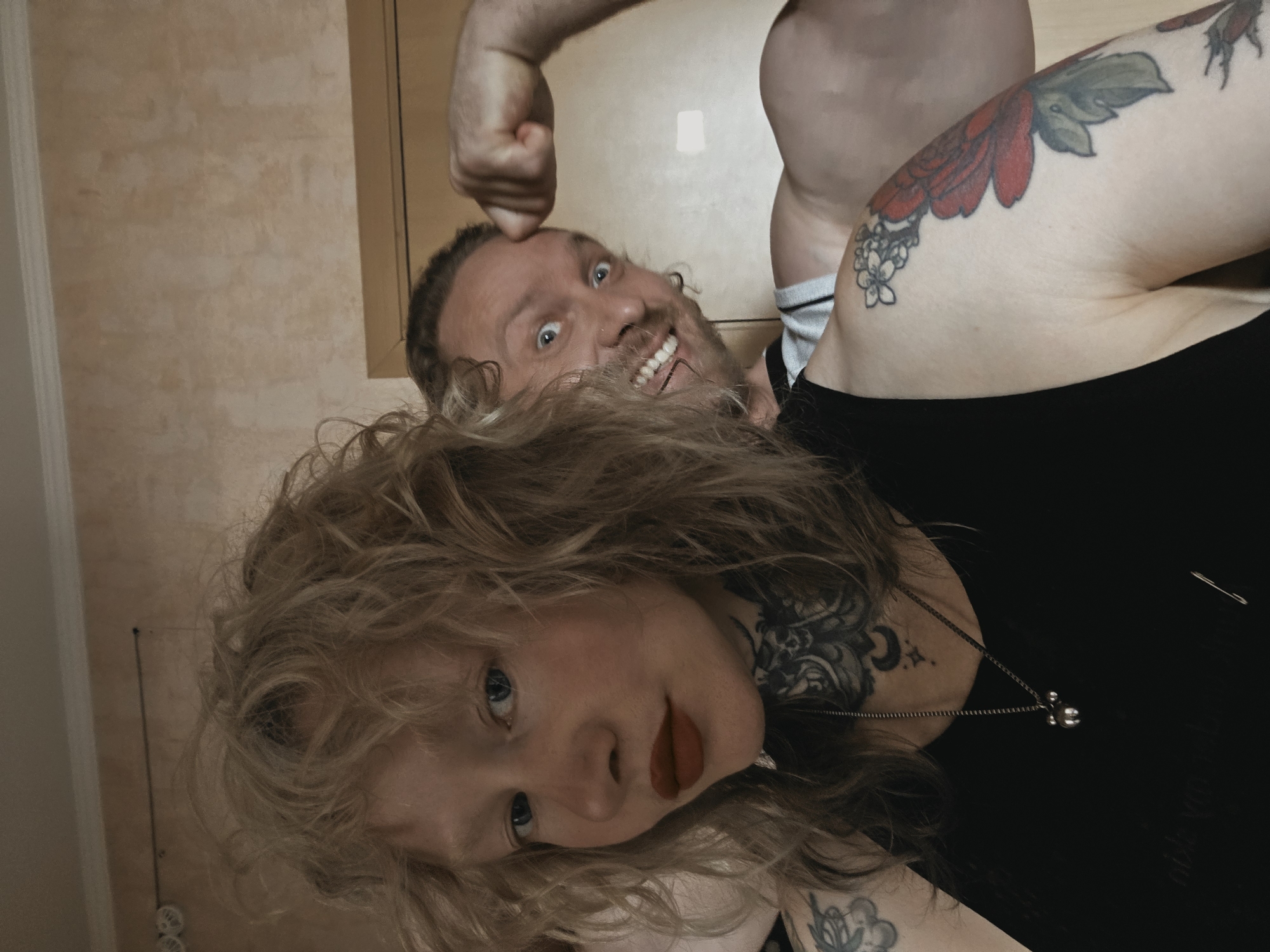 A young woman with blonde curly hair and blue eyes, wearing a black tank top and dark lipstick, is in the foreground looking at the camera. She has a tattoo on her neck and chest. Behind her, a man with blonde hair, a nose ring, and a beard is flexing his bicep and smiling widely at the camera. He is wearing a black and grey striped shirt. The background appears to be an indoor setting with a textured wall and a doorway. The lighting suggests it is daytime.