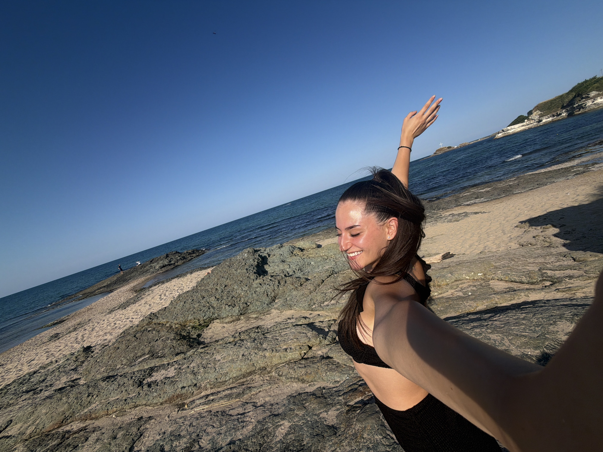 A young woman with long, dark hair pulled into a ponytail is taking a selfie on a rocky beach in Lozenets, Bulgaria. She is wearing a black bikini and smiling. Her right arm is extended towards the camera, and her left arm is raised in the air. The background shows a sandy beach, the blue sea, and a rocky coastline with green vegetation. The sky is clear and blue. There are a few people walking on the beach in the distance.