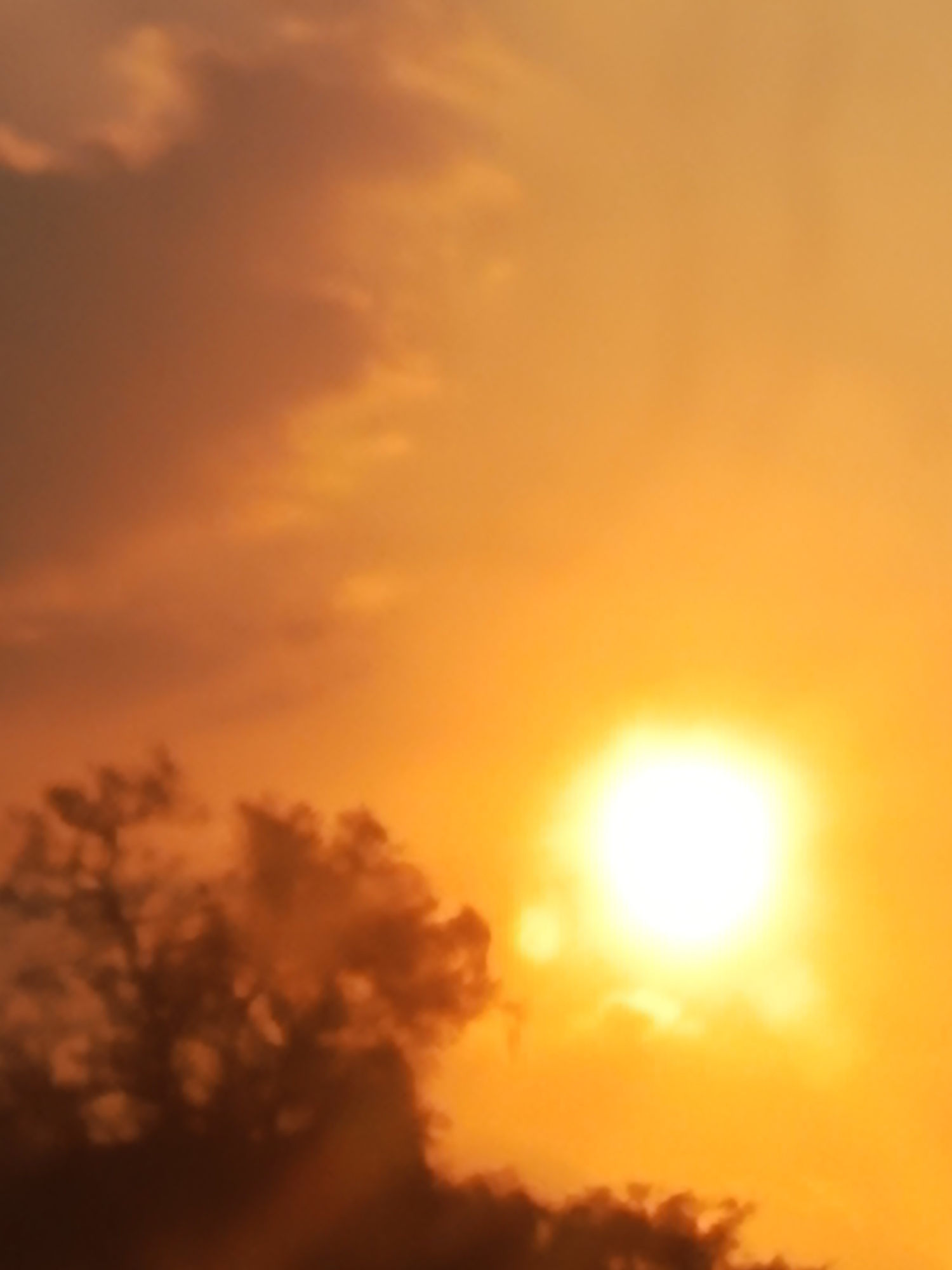 This is a photograph of a sunset over Gunung Sari, Indonesia. The sun is a bright, glowing orb in the right portion of the image, surrounded by an orange and yellow sky. Dark clouds are visible in the lower left corner, obscuring part of the sun. The image is a close-up shot, with no people or specific objects clearly identifiable besides the celestial bodies. The overall impression is of a dramatic and atmospheric sky.