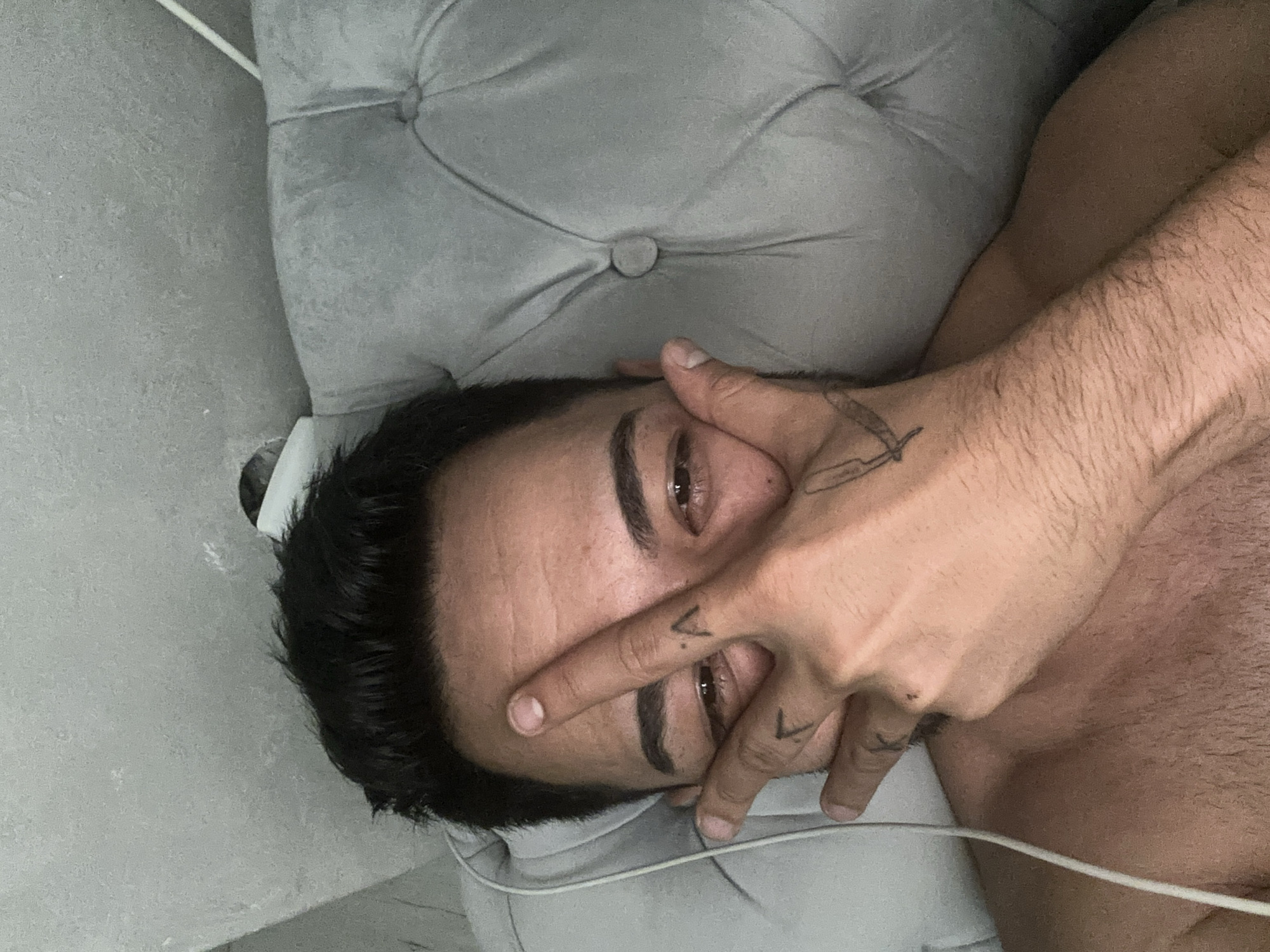 A close-up, sideways portrait captures a man lying indoors, likely in Sofia, Bulgaria. He rests his head on a light grey, tufted upholstered surface, possibly a couch or headboard. The man has dark hair and eyebrows, with visible stubble. His right hand is partially covering his face, with his index and middle fingers obscuring his forehead and upper nose, while his eyes are visible and directed towards the camera.
Distinctive tattoos are present on his hand: a razor blade design on the back of his hand/wrist, and two small V-shaped symbols on the sides of his index and middle fingers. His bare shoulder and arm, showing body hair, are visible on the right, suggesting he is shirtless. A thin white cable runs diagonally across the lower-right portion of the image. The soft indoor lighting provides a gentle ambiance. The man's visible eyes convey a relaxed or contemplative mood, despite his hand partially obscuring his expression.