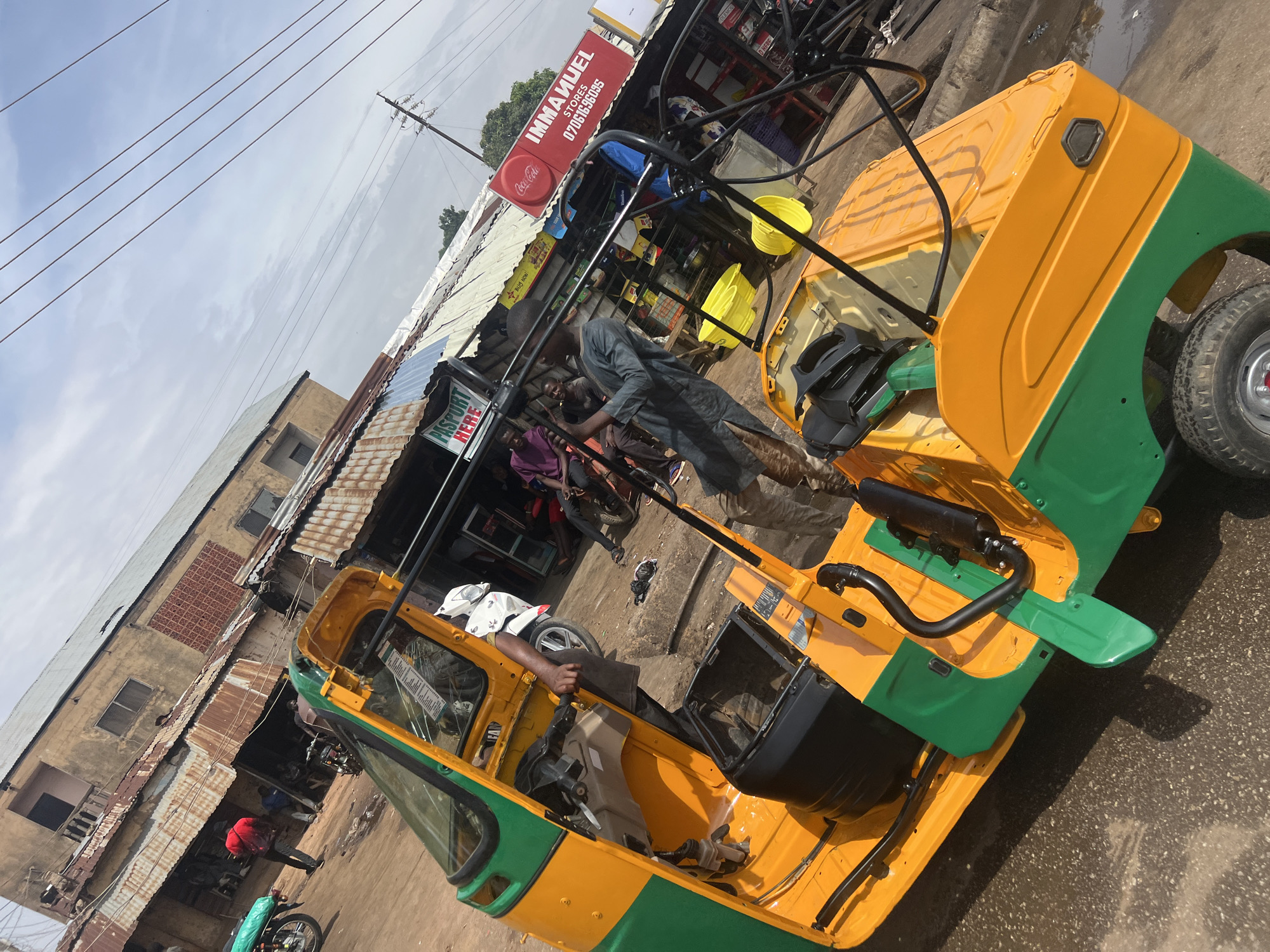 The image captures a vibrant street scene in Gura Top, Nigeria, featuring a brightly painted yellow and green tricycle (known as a keke napep) that appears to be undergoing assembly or customization. The tricycle is positioned prominently, showing its newly painted body with several internal components yet to be installed, such as seats and handlebars. A black exhaust pipe is visible at the rear.
Several individuals are present. One man wearing a white shirt and dark trousers is seated inside the tricycle's front compartment, his hand resting on what will be the steering area. Another man in a dark grey outfit stands near the front of the tricycle, observing or working on it. In the background, other people are visible near a row of shops and buildings.
The setting consists of old, somewhat weathered buildings with corrugated metal roofs, indicative of a commercial area. Power lines crisscross the sky above. Various objects are scattered around, including yellow buckets near the tricycle.
Notable signs are visible: "IMMANUEL STORES" with a phone number "07061636095" and a Coca-Cola logo are displayed on a red signboard above one of the shops. Another smaller sign reads "PASSPORT HERE."
The scene is lit by diffused daylight, suggesting a cloudy or partly cloudy day. The ground appears rough and unpaved. The overall impression is one of everyday activity in a bustling local marketplace or service area, centered around the preparation of a new or refurbished public transport vehicle.