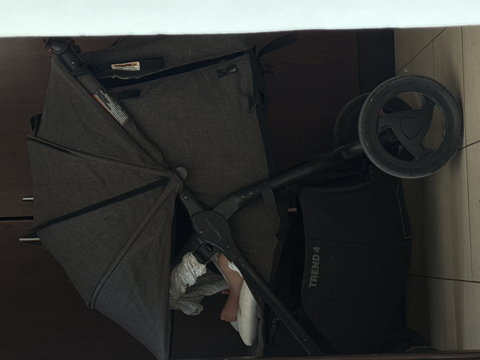 A child's foot is visible inside a dark gray stroller, which is positioned in an indoor setting with tiled flooring. The stroller has a canopy, a storage basket labeled "TREND 4," and a prominent wheel. The background appears to be a wall or closet, suggesting the stroller is not in use outdoors or in a public space. The overall lighting is dim. The image was captured in Dubai, United Arab Emirates.