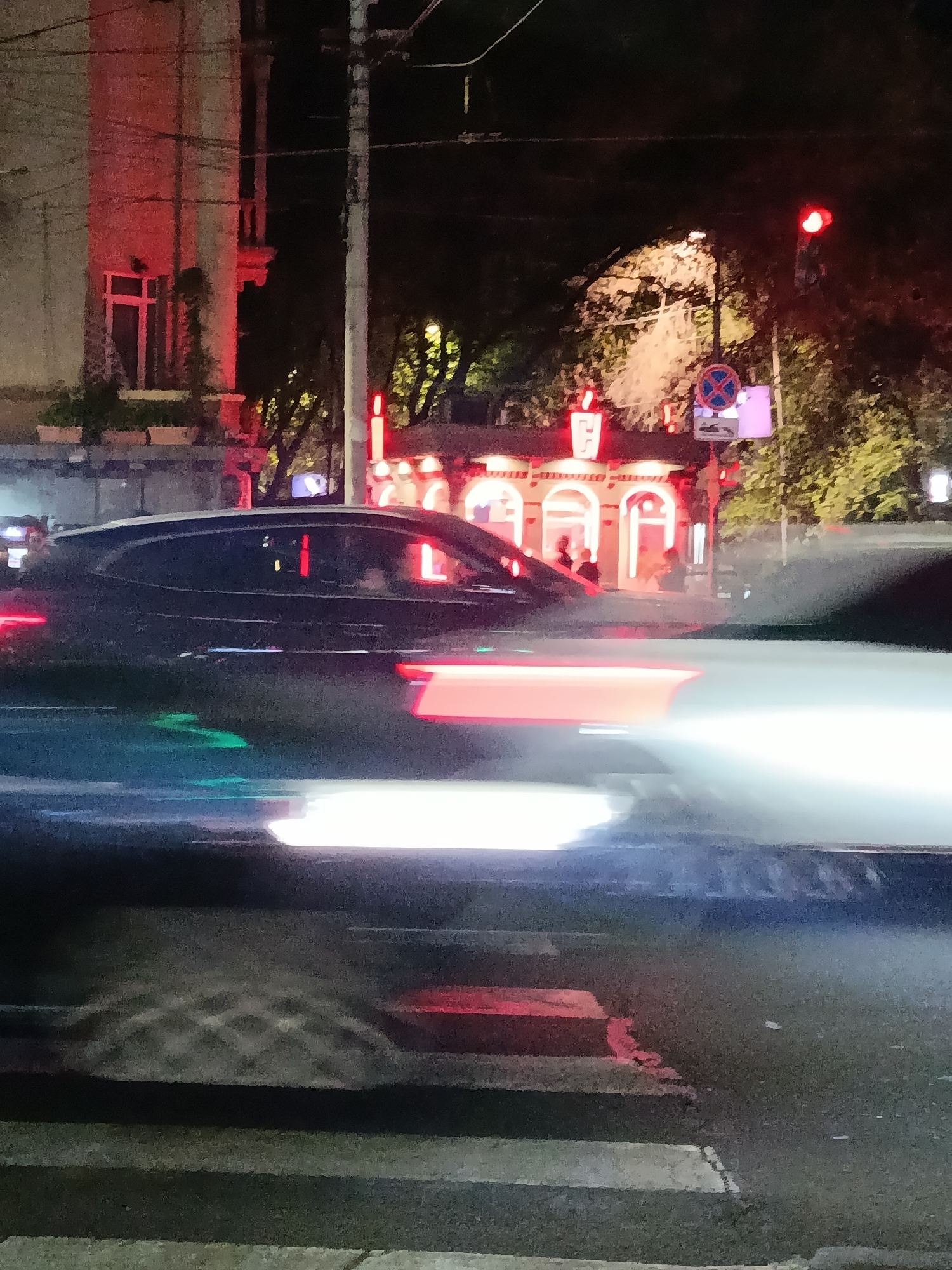 At night in Yerevan, Armenia, the scene depicts a street intersection with blurred vehicle lights. A building with red lighting features is visible behind the cars. Pedestrian traffic appears to be light, with individuals standing in or near the illuminated structure, potentially a building or a bus station. A traffic light emitting red light stands nearby. A no-parking sign is also visible in the street. A building with red accents is also seen on the left. The ground is marked by white stripes of a crosswalk.