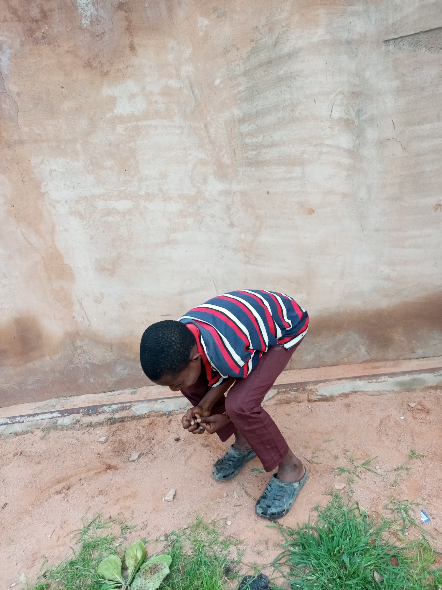 A young boy wearing a striped t-shirt and maroon pants is crouched down on sandy ground in front of a textured, light-brown wall. He appears to be examining something in his hands, which are clasped together near the ground. The wall shows signs of age and wear with cracks and discoloration. The ground is a mix of sand and sparse green vegetation. The scene is outdoors, likely in a casual or rural setting within the city of Tempure, Nigeria.