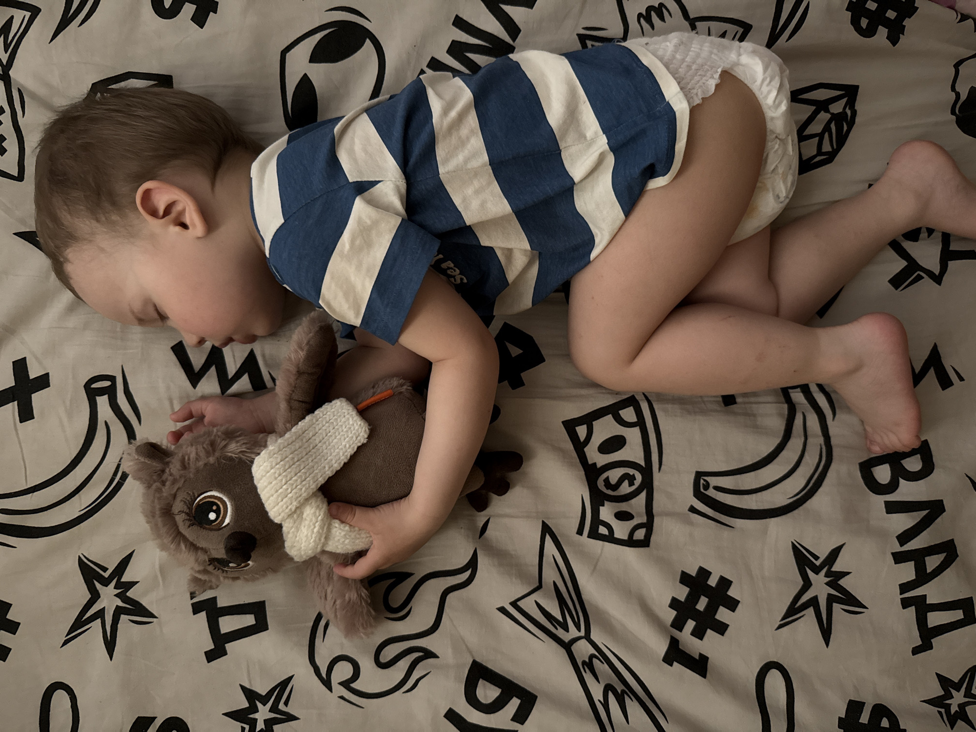 A child sleeps on a patterned bedsheet, hugging a stuffed animal. The child is wearing a blue and white striped shirt and a diaper. The bedsheet has a black and white pattern of bananas, money signs, stars, and the text "ВЛАД" and "#1". The location is unknown.