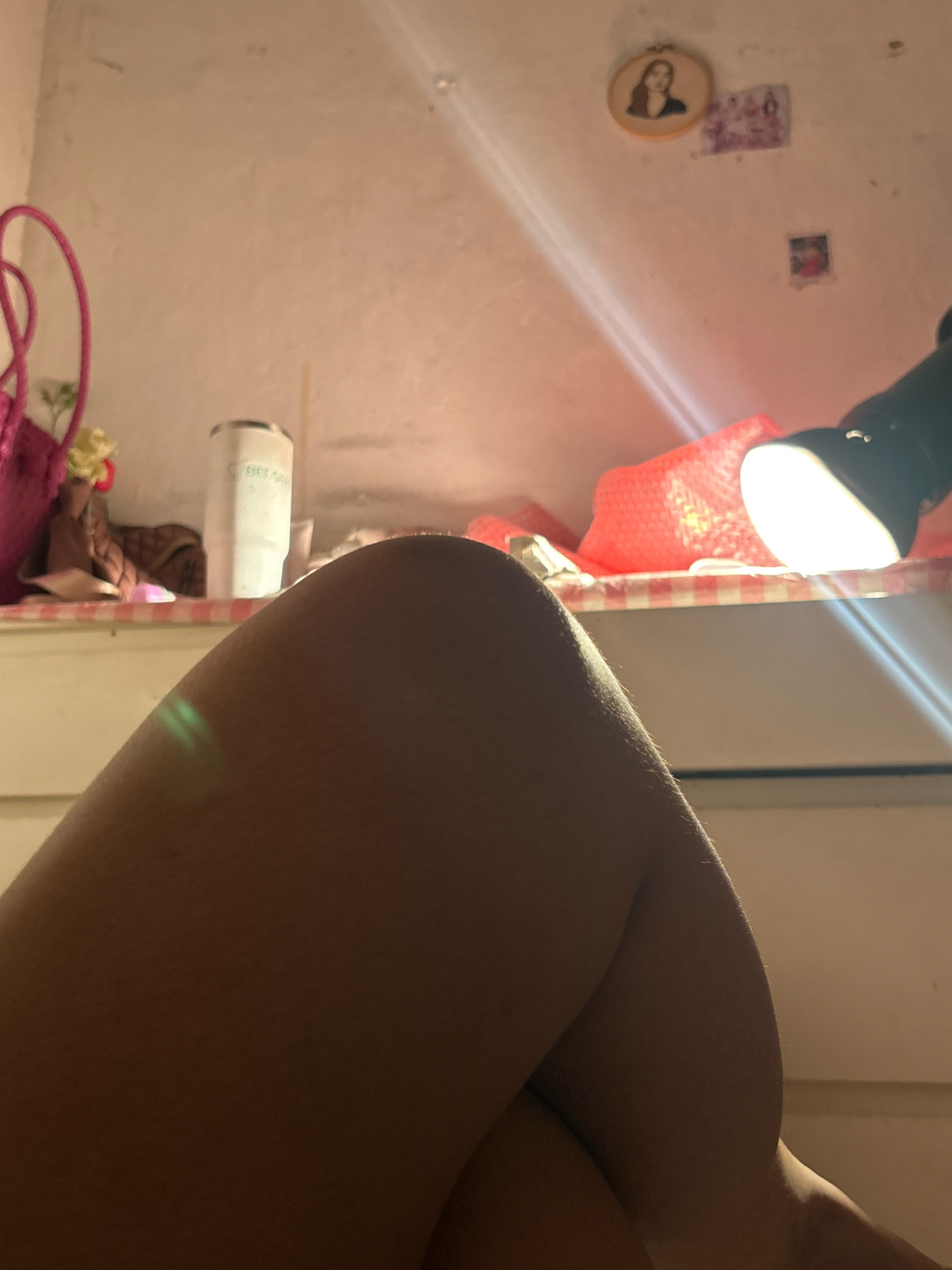 The image shows a close-up of a person's legs, which are crossed. The legs are illuminated by a bright light source, possibly a desk lamp, that creates strong highlights and shadows. In the background, there is a shelf with various items, including a white tumbler, a pink bag, and some decorative elements on the wall, such as framed pictures. The environment appears to be indoors, likely a bedroom or a study area, with a warm, somewhat dim lighting overall, except for the direct light on the legs and the lamp. There is no visible indication of the specific location in North Jakarta, Indonesia.