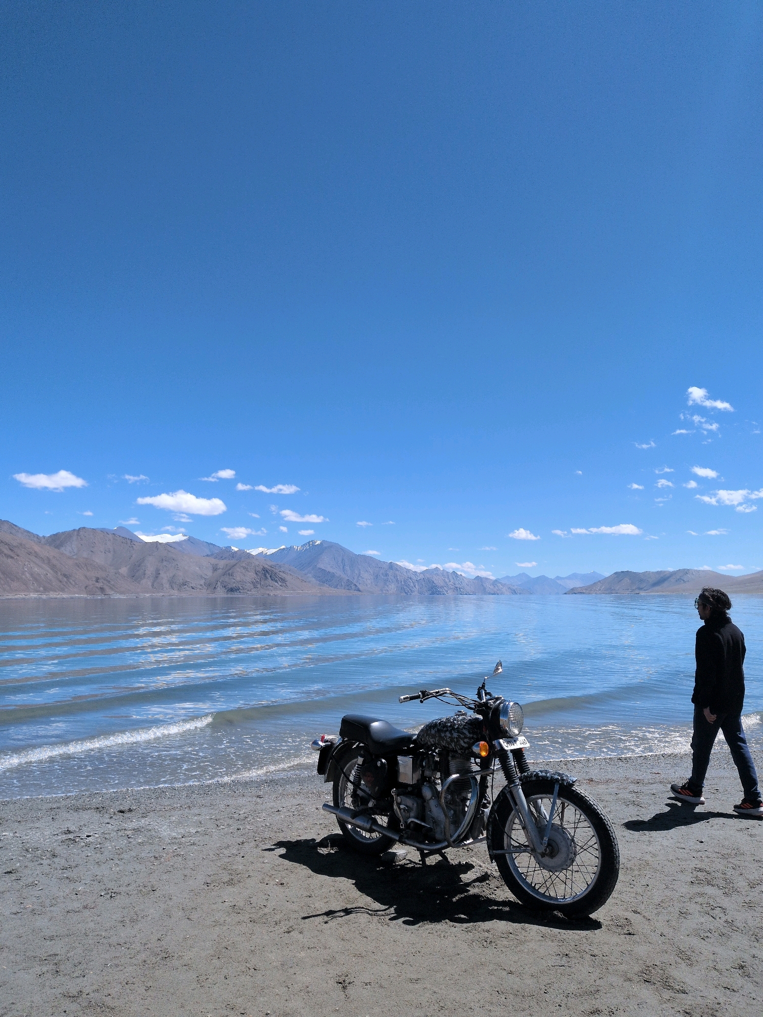 A person walks away from a parked motorcycle on a sandy shore next to a large body of water. The person is wearing a black hooded jacket and dark pants with athletic shoes. The motorcycle is dark-colored and appears to be a vintage model. The body of water is calm with gentle ripples and stretches out towards a range of mountains in the distance. The sky is clear blue with a few scattered white clouds. The scene is brightly lit by daylight. The location is the Pangong Tso lake in Ladakh, India, a high-altitude saline lake known for its stunning blue waters and surrounding mountain landscape.