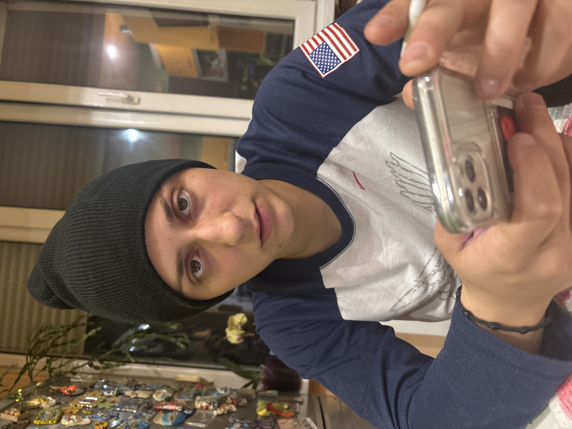A person wearing a black beanie and a long-sleeved shirt with an American flag patch on the sleeve is holding a smartphone in a clear case. The person is looking at the camera. In the background, there is a table covered with an array of souvenirs, potentially fridge magnets. A houseplant is also visible on the table. There are closed blinds or a window in the background. The exact location within Sofia, Bulgaria is not discernible from the image alone.