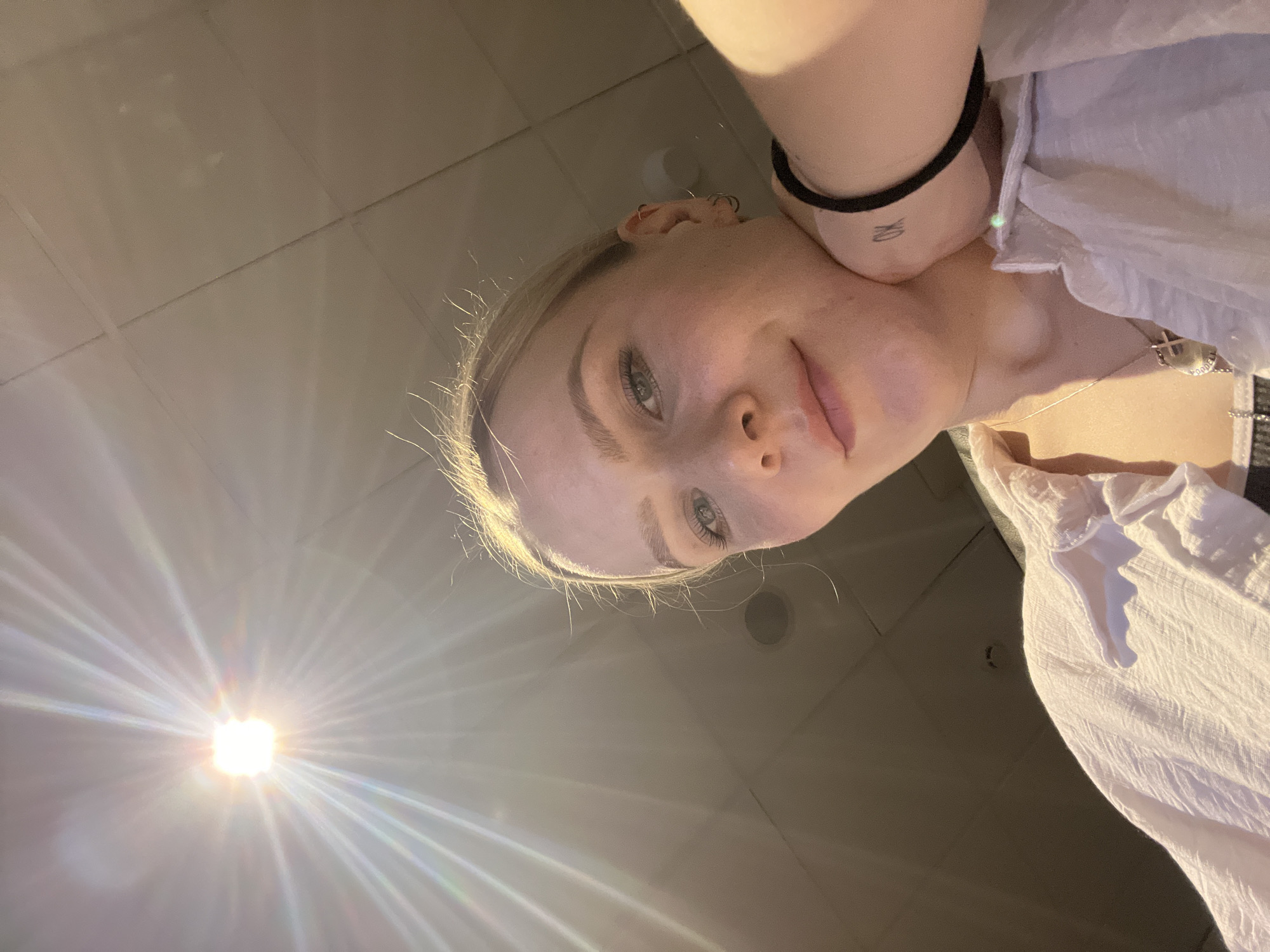 The image presents a low-angle view of a young woman with fair skin and light blonde hair. She is wearing a white, textured shirt. Her left wrist features a black bracelet and a small tattoo. She is looking downwards towards the camera. The background consists of a lightly-colored, tiled ceiling with recessed lighting. A bright light source is visible in the lower-left corner of the image. There is no evidence to suggest the location is in Khimki, Russia.