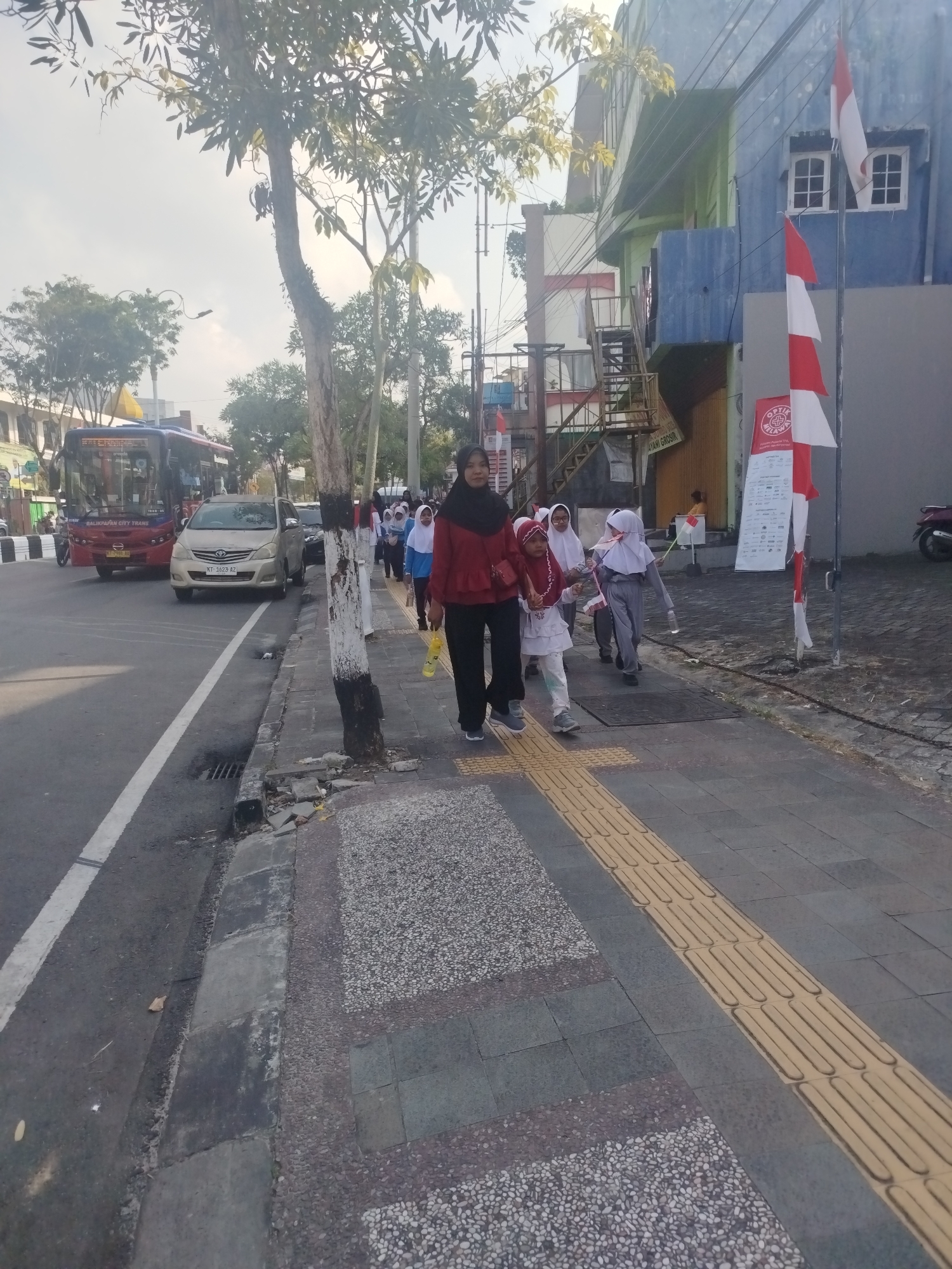The image shows a street scene in Balikpapan, Indonesia, with a sidewalk on the right and a road on the left. A group of people, mostly young girls in hijabs, are walking on the sidewalk. One woman in a red hijab and black pants is leading the group. A bus and a car are visible on the road. The buildings on the right are painted in blue and green, with Indonesian flags displayed on a pole. The sky is overcast, and there are trees and street lights visible. The sidewalk has textured paving for the visually impaired.