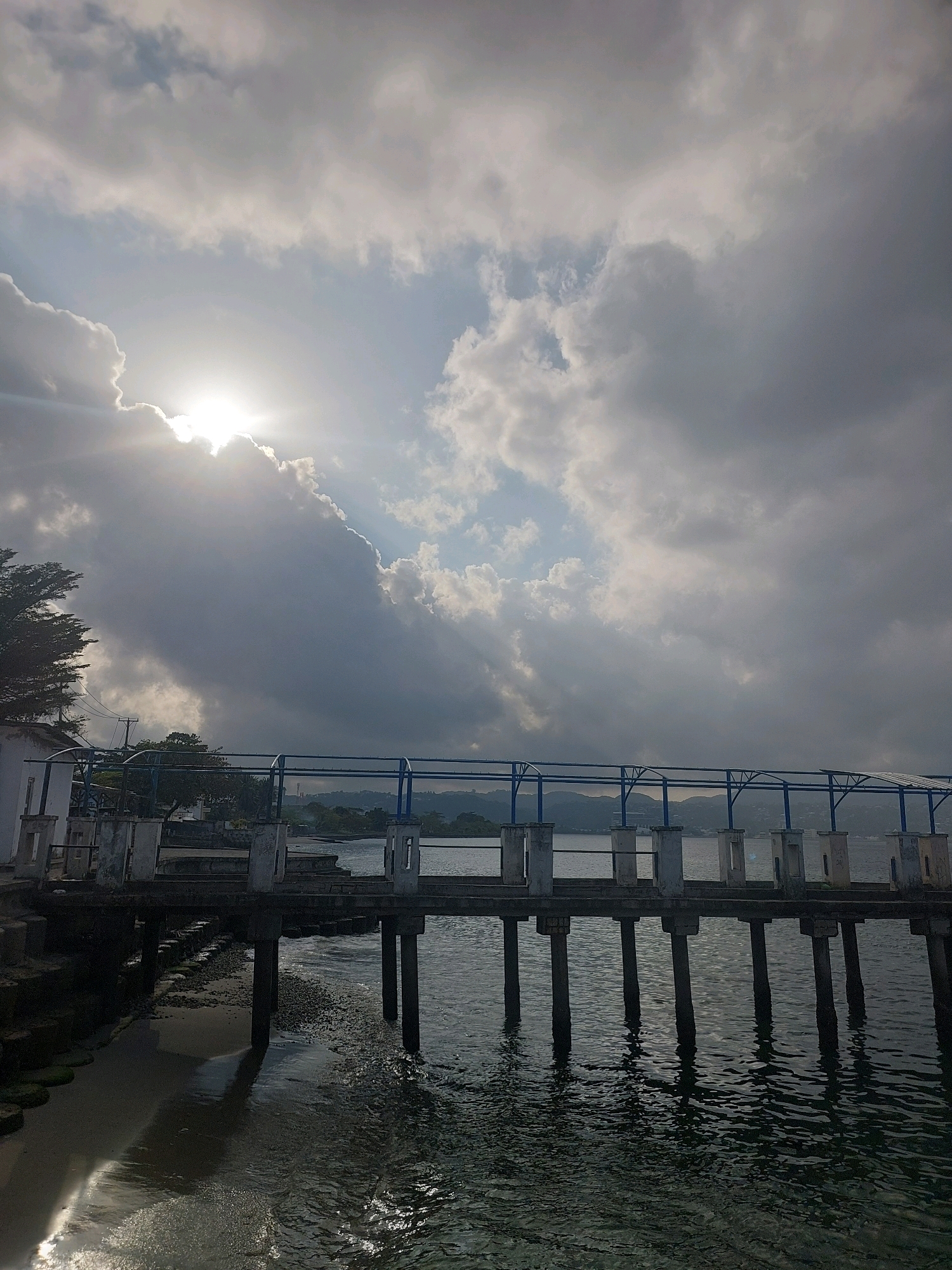 The image depicts a pier extending over calm water, with a cloudy sky illuminated by the sun peeking through the clouds on the left side. The pier is constructed with concrete pillars and a railing. The foreground shows the edge of the water with a sandy shoreline. In the background, there are hills and trees. The scene appears to be set in a coastal area, likely the city of Ambon, Indonesia, given the context. There are no people visible in the image.