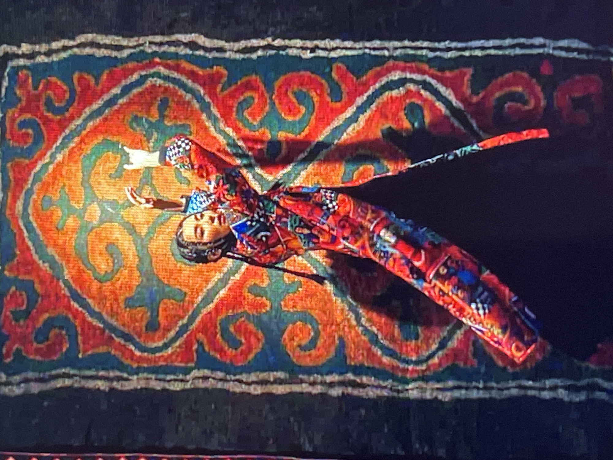 A woman in a vibrant, intricately patterned robe dances in front of a large, colorful tapestry. The robe features a striking mix of red, blue, and white geometric designs, with black and white checkered accents on the sleeves. Her hair is styled in braids that fall over her shoulders. Her arms are raised, with her right hand extended upwards in a gesture of movement. The tapestry behind her is dominated by bold orange, red, and teal swirling patterns, with a dark background. The lighting creates a shadow of the woman on the tapestry. This scene likely takes place in a performance setting or cultural exhibition in Bishkek, Kyrgyzstan, possibly at the Kyrgyz National Academic Opera and Ballet Theater or a similar venue showcasing traditional arts.
