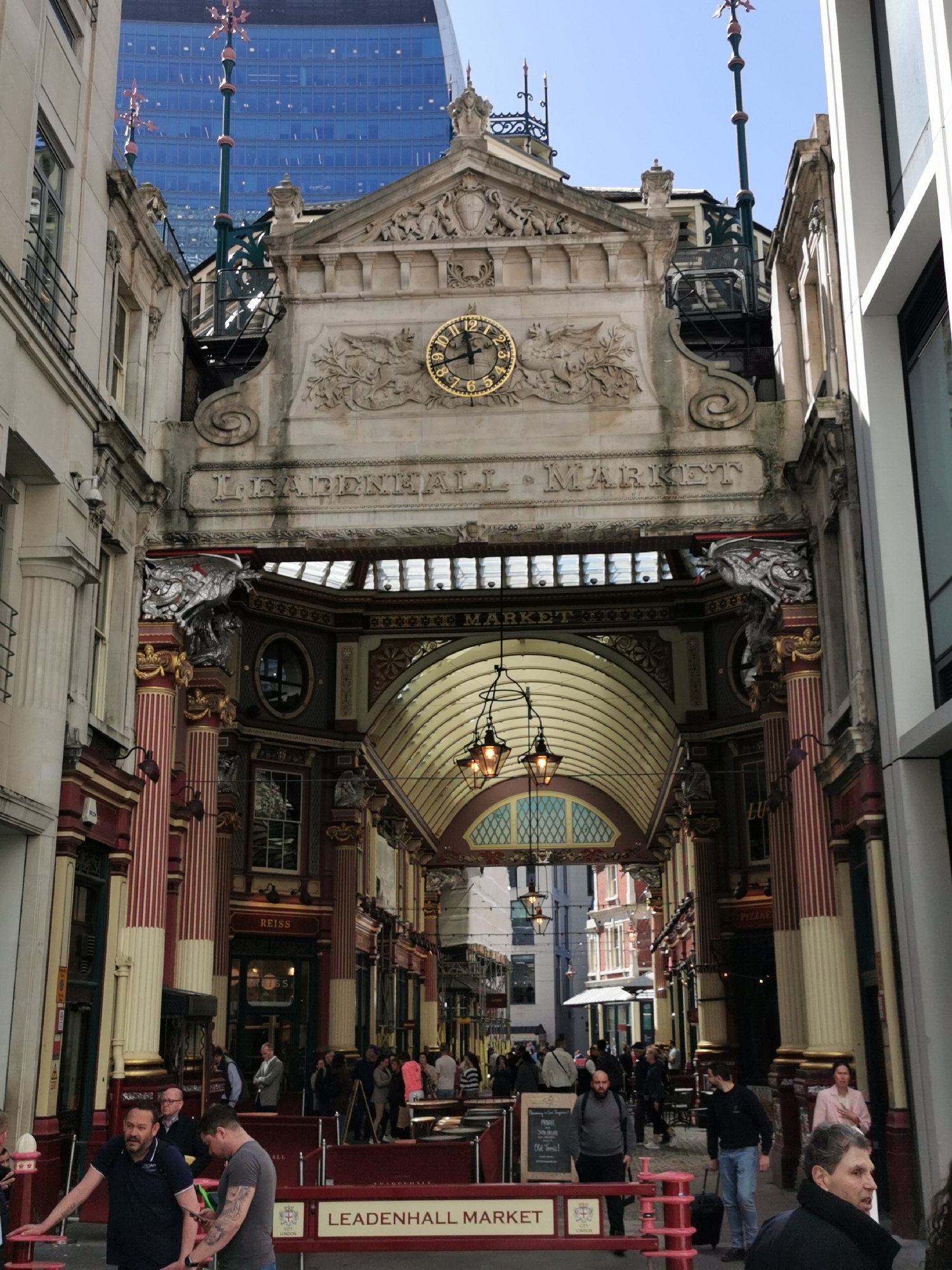 The image presents an ornate entrance to Leadenhall Market in London, United Kingdom, looking down a covered arcade on a clear, sunny day. The foreground is dominated by a grand, classical stone facade featuring a prominent golden clock, whose hands indicate approximately 12:45. Above the clock, a richly detailed pediment and carved architectural decorations, including a winged creature motif, are visible. Tall, decorative spires rise from the top of the building. In stark contrast, a modern blue-glass skyscraper towers in the background, peeking over the historic structure.
Below the main facade, the entrance to the market arcade is flanked by intricate silver dragon sculptures. The arcade itself boasts a vaulted glass ceiling, allowing ample natural light, and is illuminated by elegant hanging lamps. Along the sides, shops and establishments are housed behind facades with distinctive red and gold-striped columns. Visible text includes "LEADENHALL MARKET" on a signpost in the lower foreground, "REISS" on one shopfront, and "PIZZAE" further down the arcade, indicating a pizza vendor.
Numerous people are seen throughout the market, casually dressed, walking, standing, and engaging in conversations, contributing to a lively, bustling atmosphere. Red barriers help guide pedestrian traffic. The overall scene captures a vibrant urban environment where historical architecture and modern development coexist seamlessly under a bright blue sky, suggesting pleasant daytime weather.