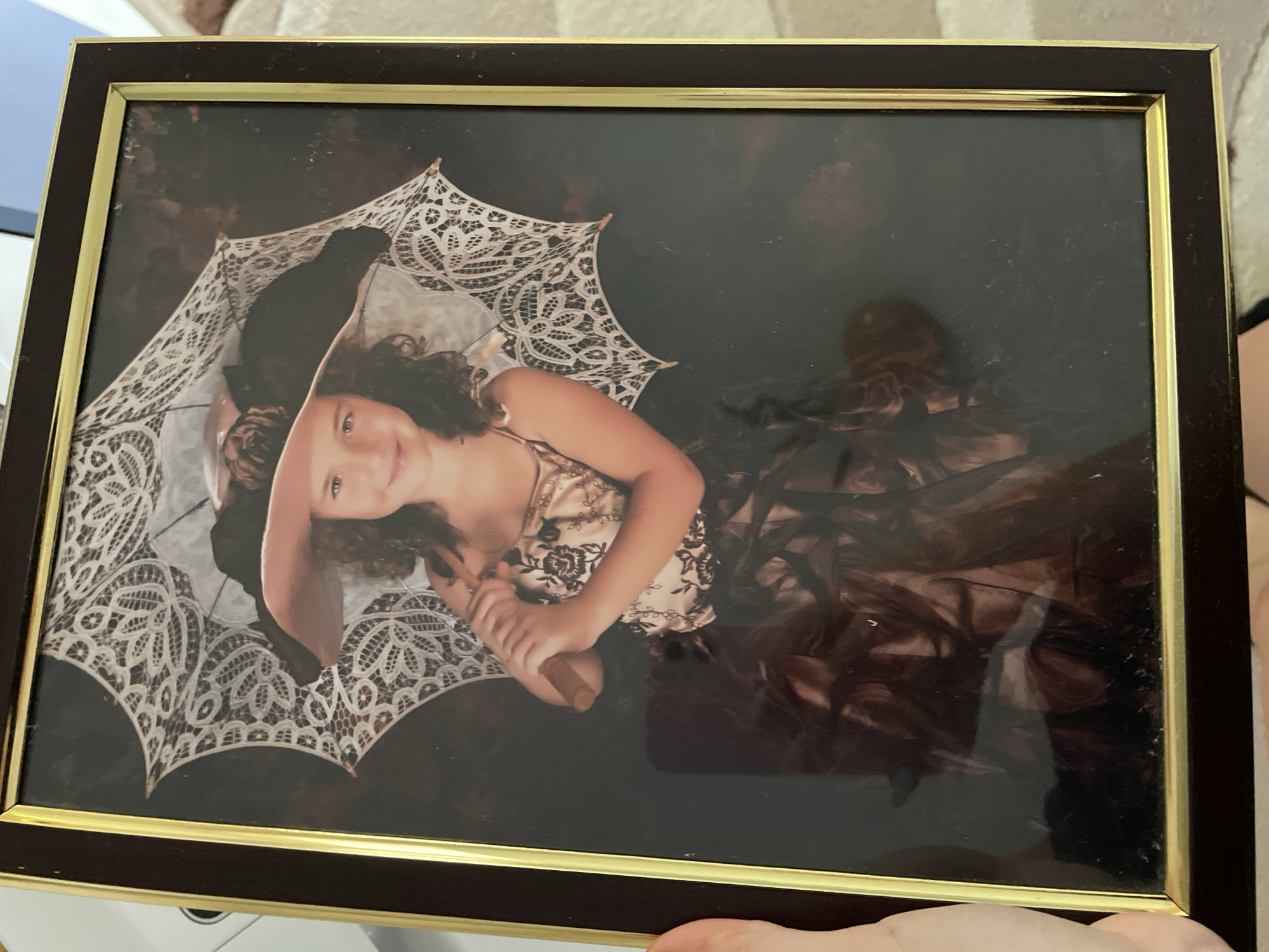 A framed portrait photograph shows a young girl in a costume. The girl is smiling and holding a lace umbrella. The background is dark and appears to be a studio setting. The photograph is in a dark brown frame with a gold inner border. The image does not contain any information about its location in Zlatograd, Bulgaria.