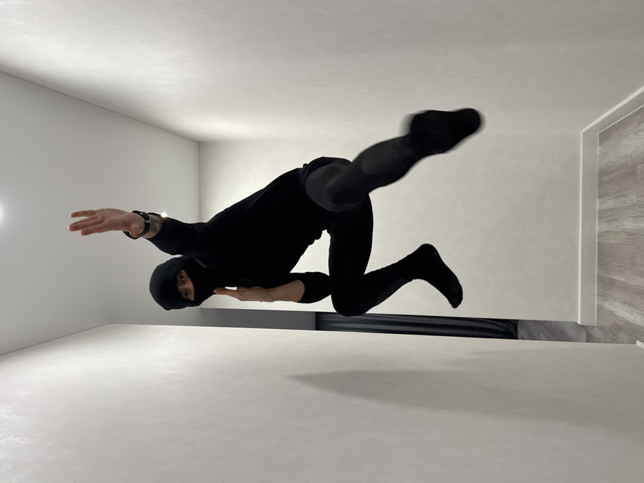 A person dressed in all black, including a balaclava, is suspended horizontally in a room with white walls, floor, and ceiling. The individual is positioned mid-air, extending one arm and a leg outward, creating a dynamic, superhero-like pose. A dark horizontal line runs across the middle of the image, possibly indicating a support or visual effect. The room appears empty except for the person and this dark line.
