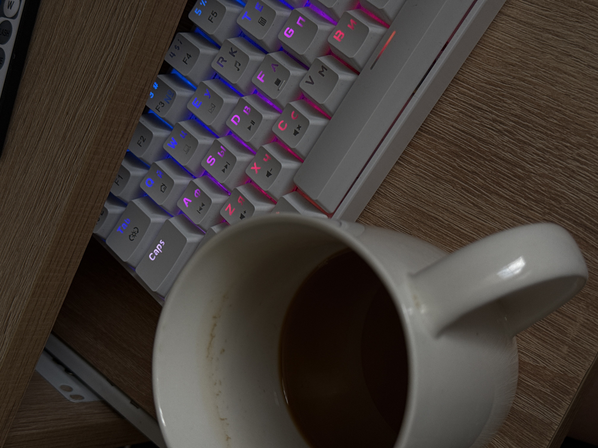 The image shows a white coffee mug filled with dark liquid, placed on a wooden desk next to a white mechanical keyboard. The keyboard has RGB backlighting, with keys displaying various letters and symbols in different colors. Some keys, like "Caps," "Tab," and F-keys, are visible. The desk appears to be part of a shelf or furniture unit, indicated by the wooden planks and a metal bracket visible in the background. The lighting is dim, contributing to the overall moody atmosphere of the scene. There are no people or signs of activity depicted. The location is not specified.