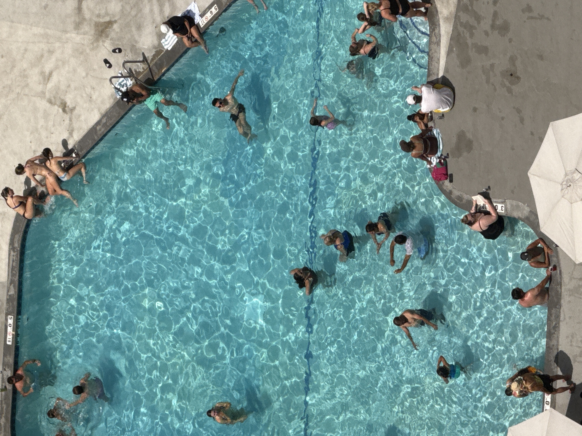 An overhead shot captures a vibrant scene at an outdoor swimming pool, likely during the daytime on a sunny day. The pool's clear blue water, illuminated by sunlight, reflects shimmering patterns on the surface. Several people are enjoying the water, some swimming, while others relax or socialize around the pool's edge.
In the upper left corner, a group of people are seated on the pool deck, some with towels. Near them, a person in a black shirt and white hat is sitting on the edge of the pool. In the water, several individuals are swimming and interacting.
Moving into the center of the image, more people are visible swimming. One person in a black swimsuit is floating on their back, while others are actively swimming or standing in the water. A few individuals are also visible near the edge of the pool, either entering or exiting the water.
Towards the right side of the frame, a person in a yellow shirt and white hat is sitting on the pool deck, and another person in a black swimsuit is reclining. Further down, a person in a white shirt is leaning against the pool's edge.
The pool has depth markers visible on its edge, with numbers like "3 0 12" and "5 0 15" indicating distances. Two white beach umbrellas are partially visible at the bottom right, suggesting a recreational area. The overall atmosphere appears relaxed and enjoyable, with people engaging in various leisure activities at the pool. The scene suggests a warm day, perfect for swimming and outdoor fun.
