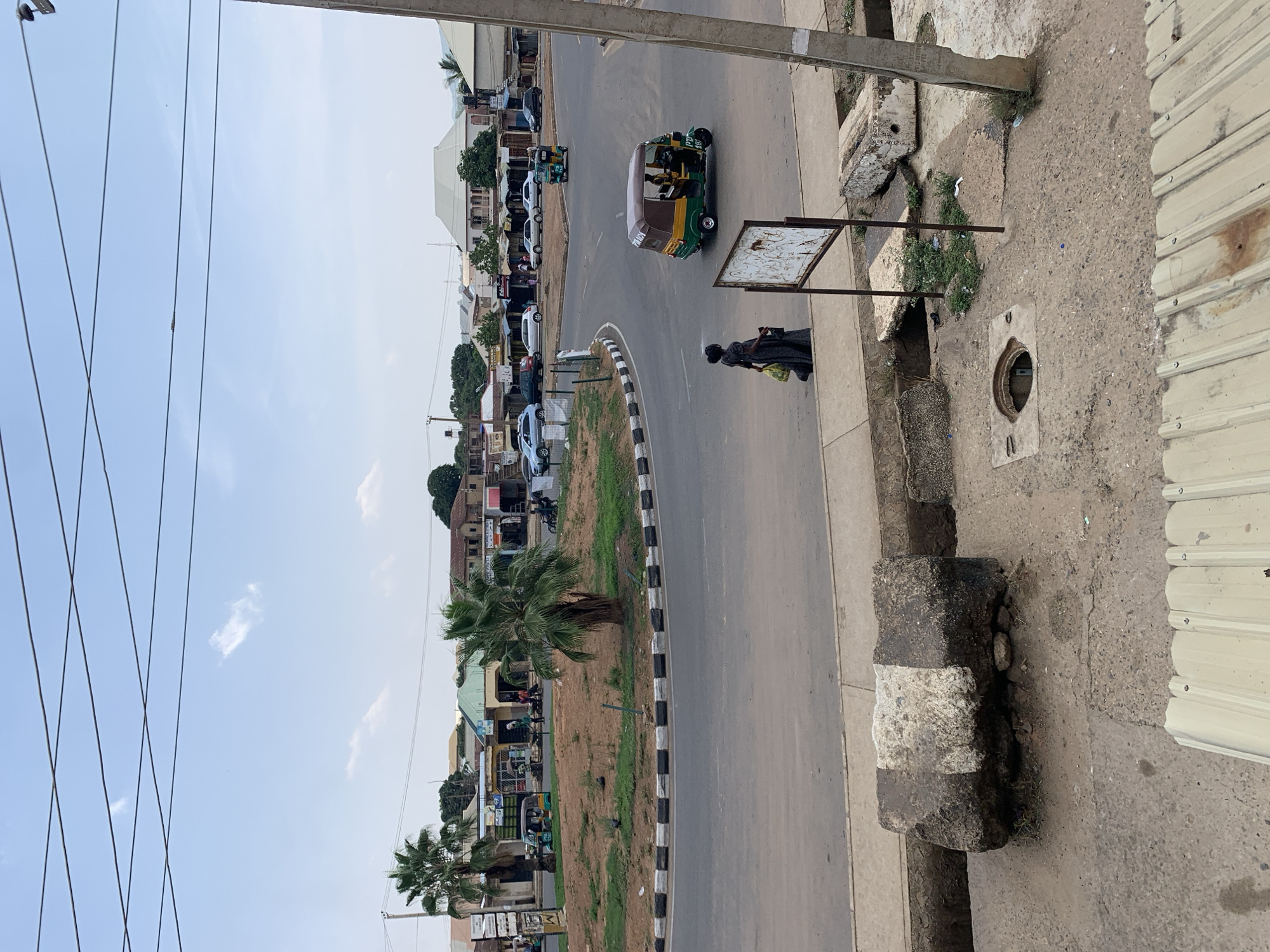 The image depicts a street scene in Rafin Par, Nigeria. The road curves, with a median strip featuring patchy grass and small palm trees bordered by black and white markings. Several cars are parked along the street's left side, adjacent to buildings. A three-wheeled vehicle is visible on the road, moving away from the viewer. A person is walking on the sidewalk on the right side of the road. An open manhole is present. Overhead, electrical wires crisscross the sky. The sky is a pale blue. The image appears to be taken from an elevated position.