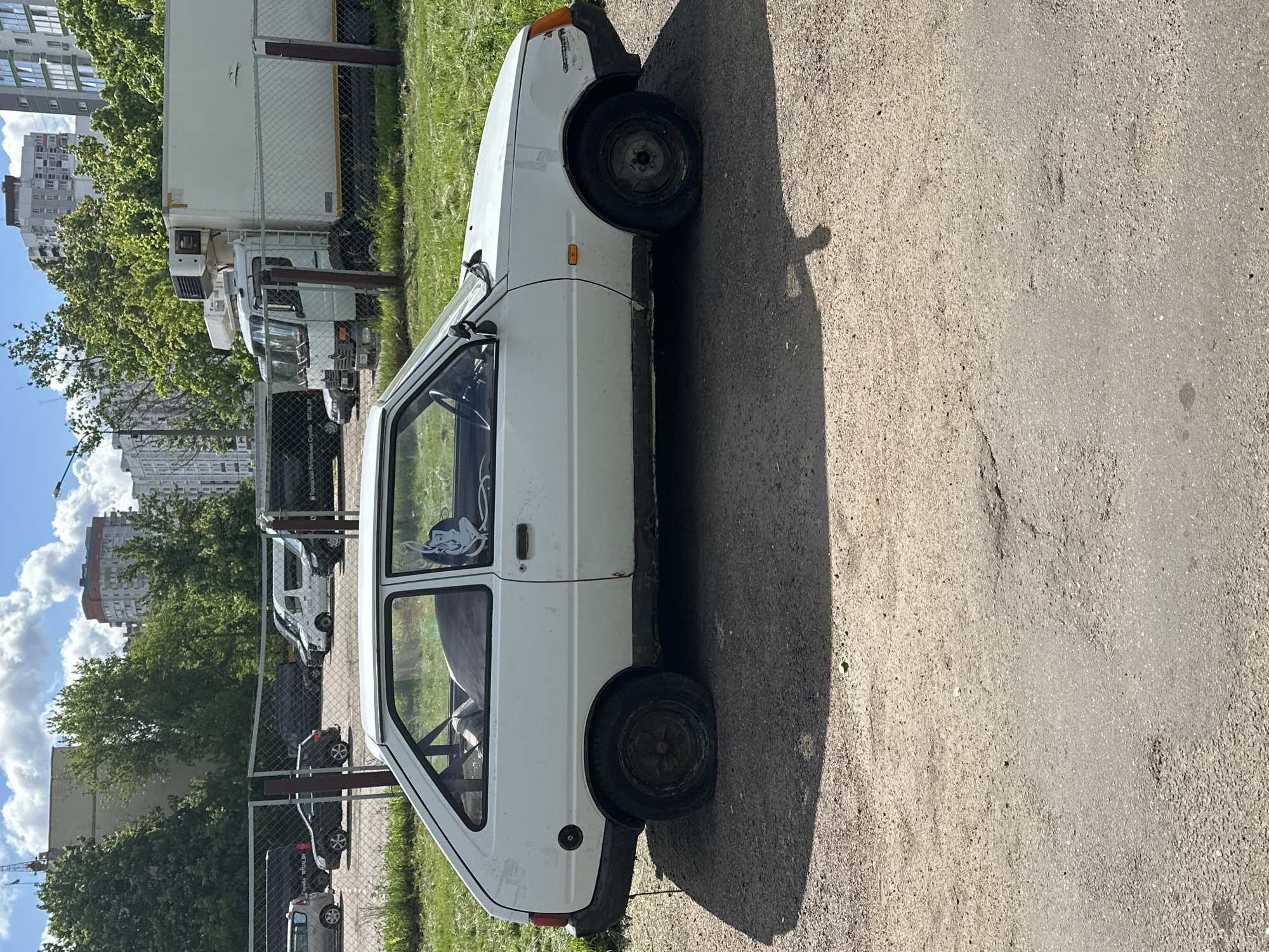A white Lada car is parked on the asphalt of a street, casting a dark shadow on the ground. Behind the car, through a chain-link fence, are several parked vans and cars. In the background are apartment buildings and trees under a partly cloudy sky. The location is Minsk, Belarus.