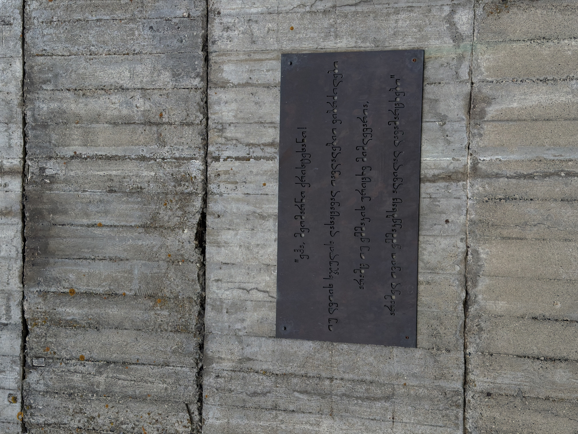 A landscape, eye-level, full shot photograph depicts a grey concrete wall with a dark grey metal sign affixed to it. The wall is made of stacked, horizontal concrete blocks with vertical seams between them. There are slight variations in texture and tone across the blocks, adding a sense of age and weathering.
The sign is rectangular and features text in Georgian script. The text is engraved or etched into the metal, creating a contrast between the dark metal and the lighter characters. The sign appears to be attached to the wall with screws or bolts, visible at the corners.
The image is clean and well-lit, capturing the texture of both the concrete and the metal sign. The location of the sign is in the city of Zemo Nichbisi, Georgia.