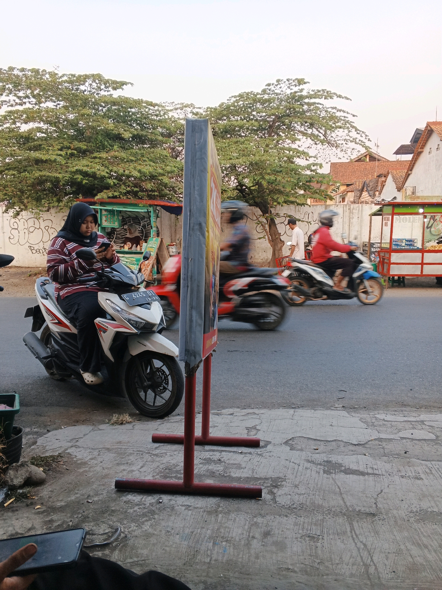 The image captures a street scene in Taman, Indonesia. In the foreground, a woman wearing a hijab and a striped shirt is seated on a white and red scooter, looking at her phone. The scooter has a license plate that reads "G 4745 DT". A blurred red scooter is visible behind her. Further down the street, two more scooters are in motion, their riders blurred due to movement. On the right side of the image, a food stall with a red and white cart displays items for sale. In the background, there are trees, buildings with tiled roofs, and a wall with graffiti. The street is paved with asphalt, and a concrete sidewalk is visible in the foreground with some debris and a potted plant. The sky is overcast.
